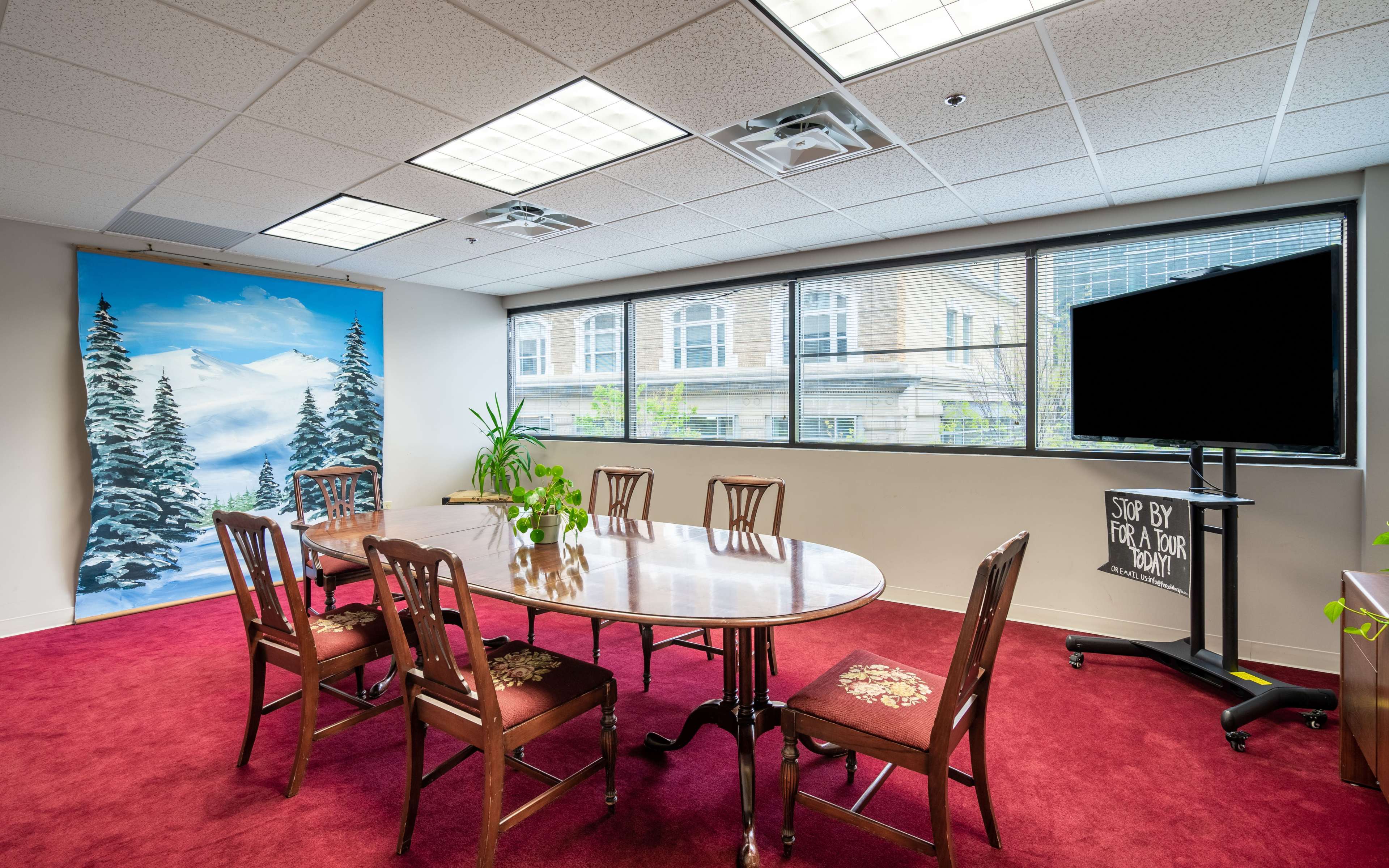 A conference room features a large wooden table surrounded by chairs, with a mountain-themed mural on one wall and a television screen on the opposite side.