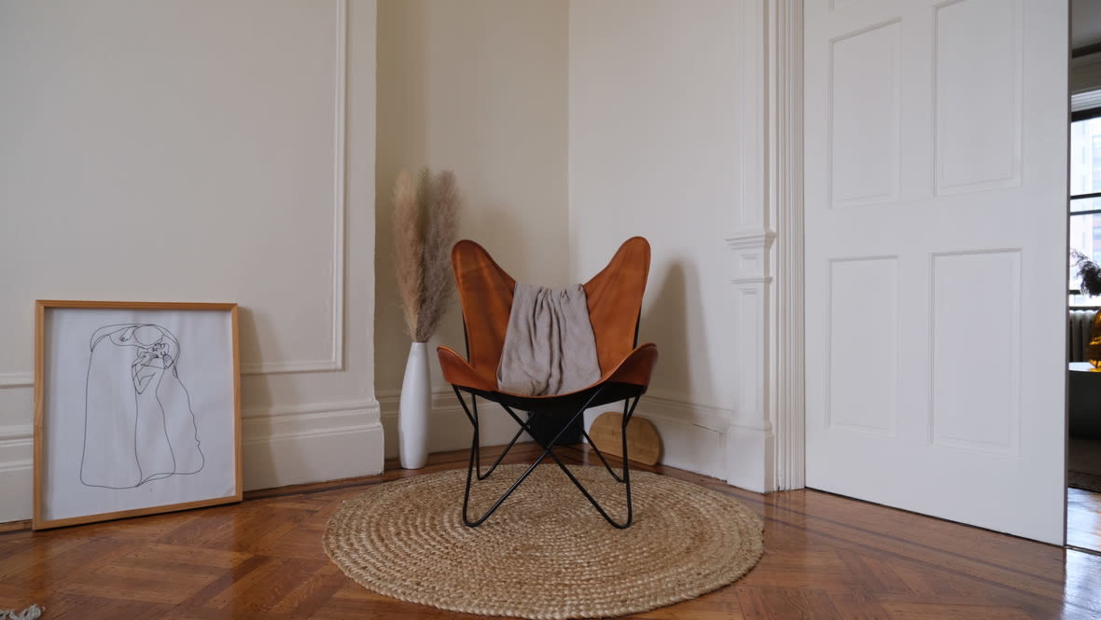 A butterfly chair with a gray draped fabric sits beside a framed line drawing and a tall vase with dried pampas grass on a circular rug in a room with light-colored walls.