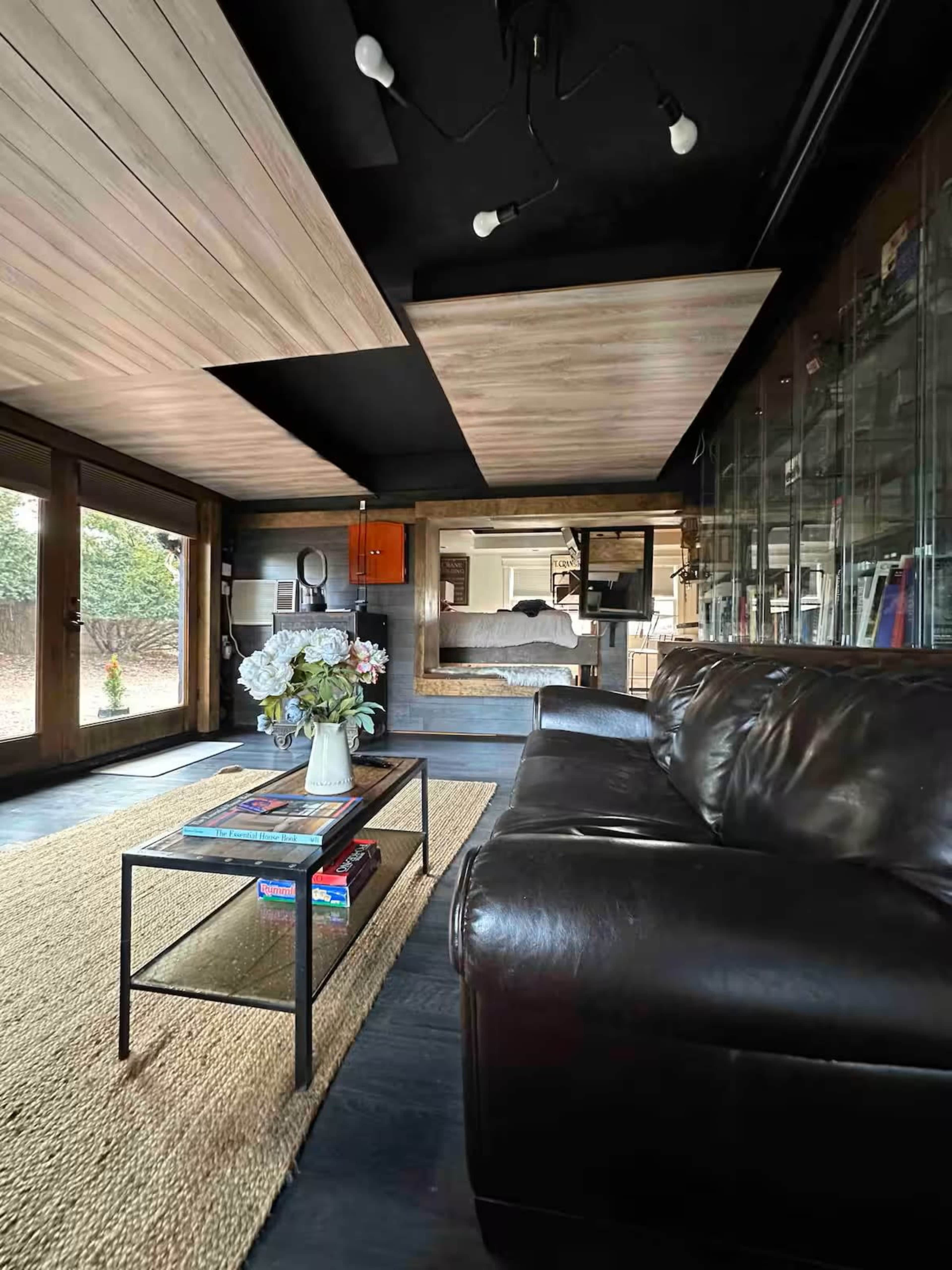 The image shows a modern living room featuring a dark leather sofa, a glass coffee table with books, and a well-lit area with large windows.