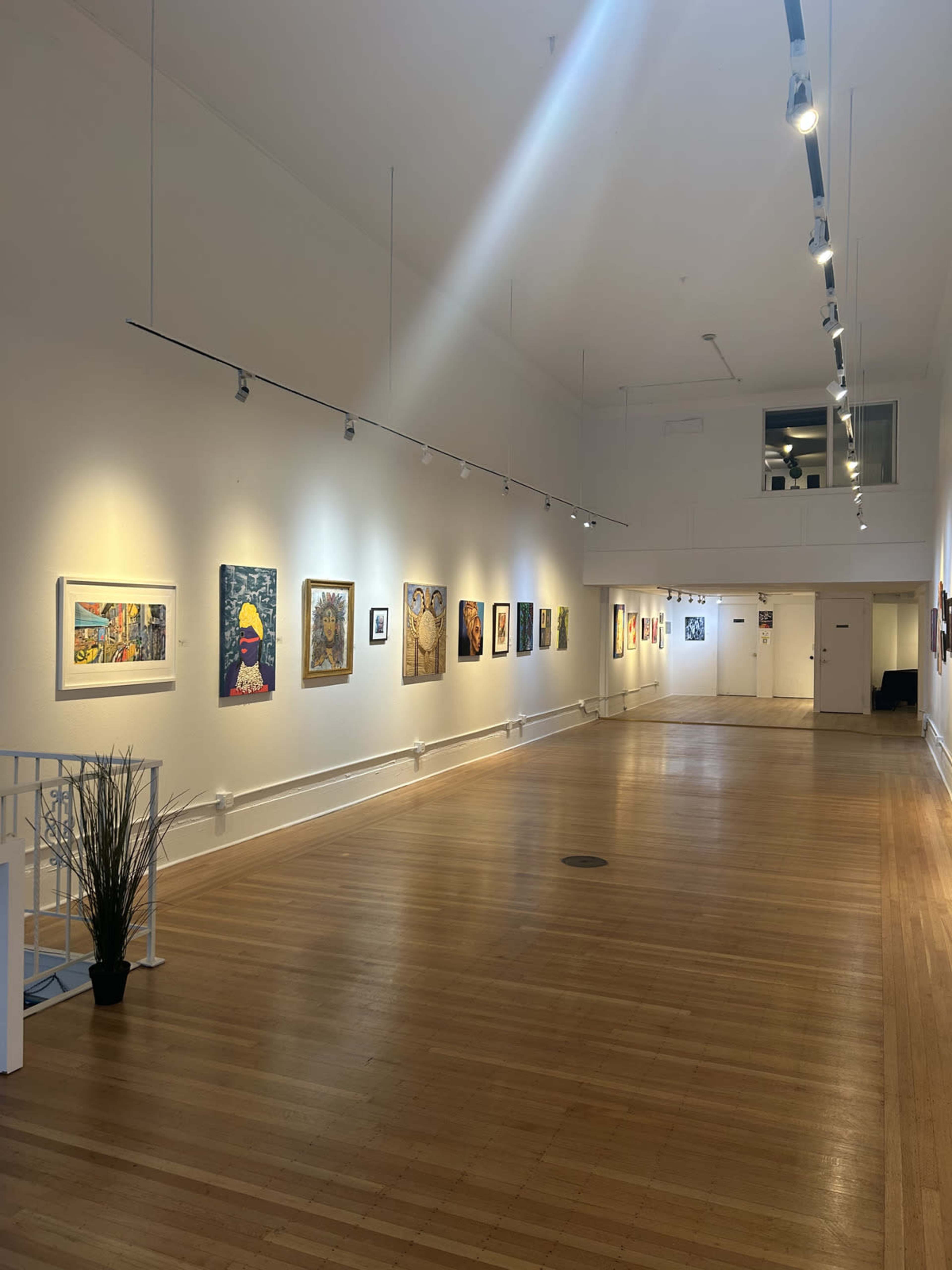 The image shows a well-lit art gallery with a long wooden floor and walls displaying various artworks.