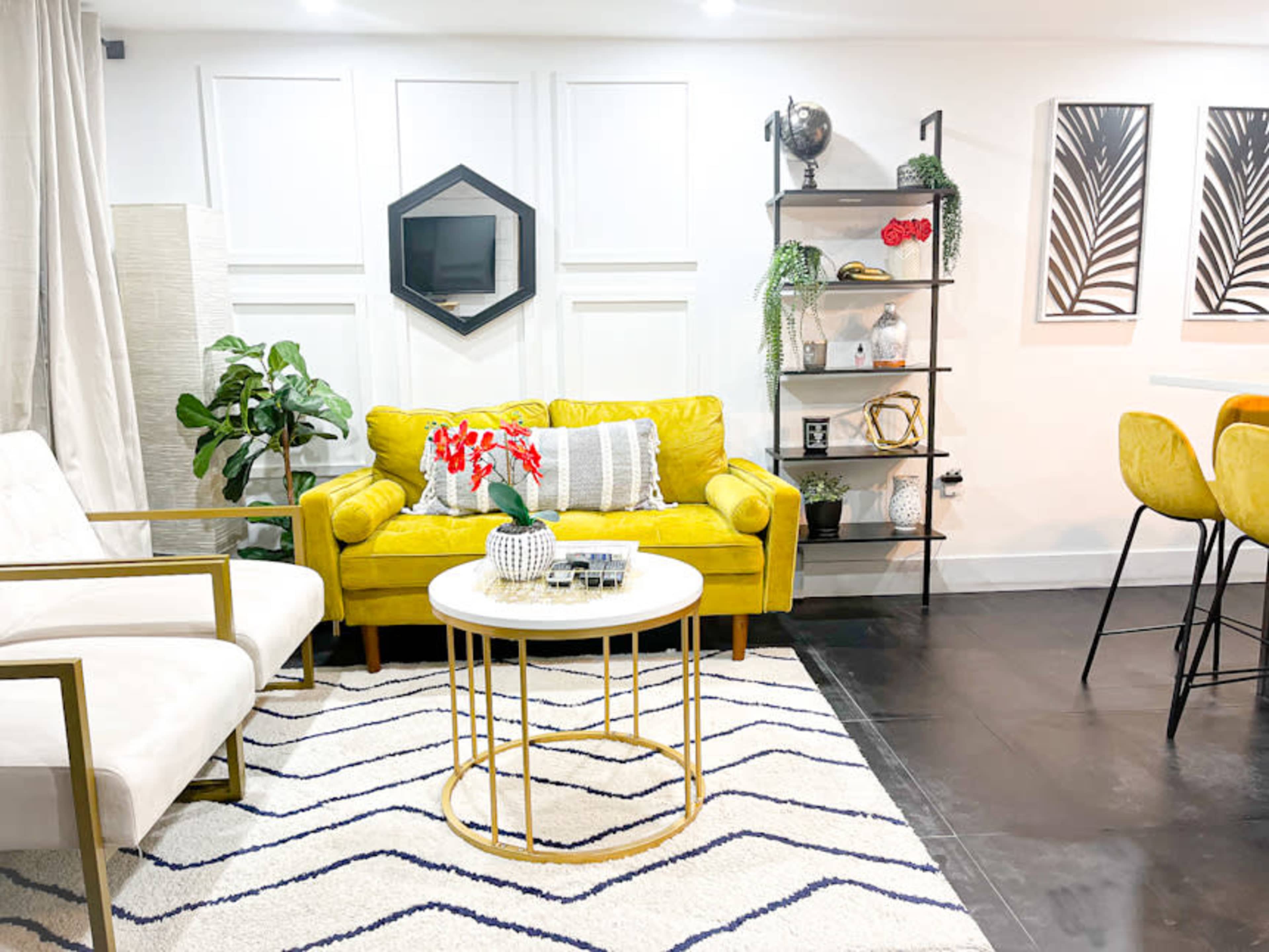 A modern living room with a yellow couch, white chairs, a round coffee table, and decorative shelving against a white wall.