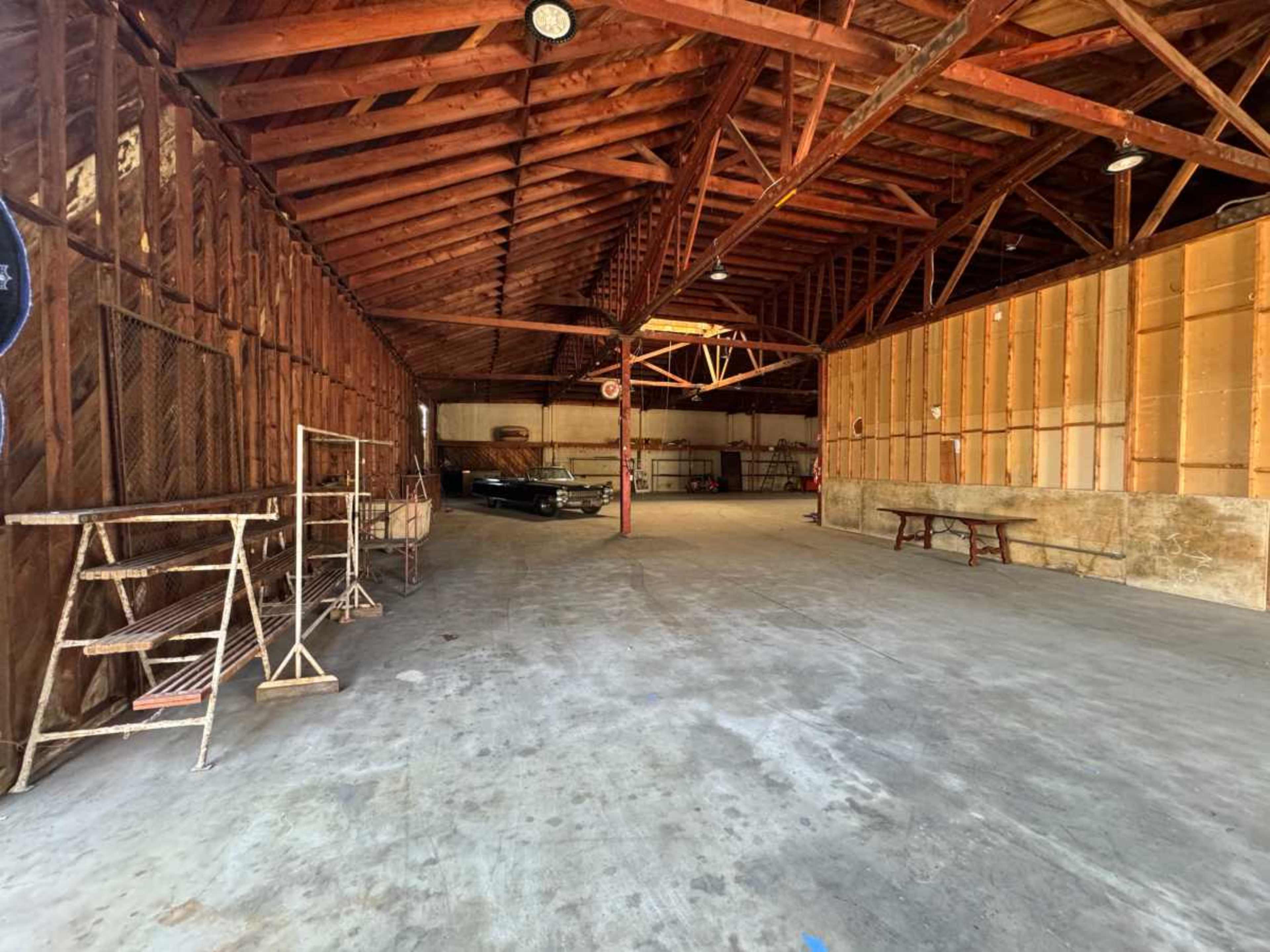 The image shows a spacious, empty barn with exposed wooden beams and concrete flooring, featuring a few pieces of equipment along the walls.