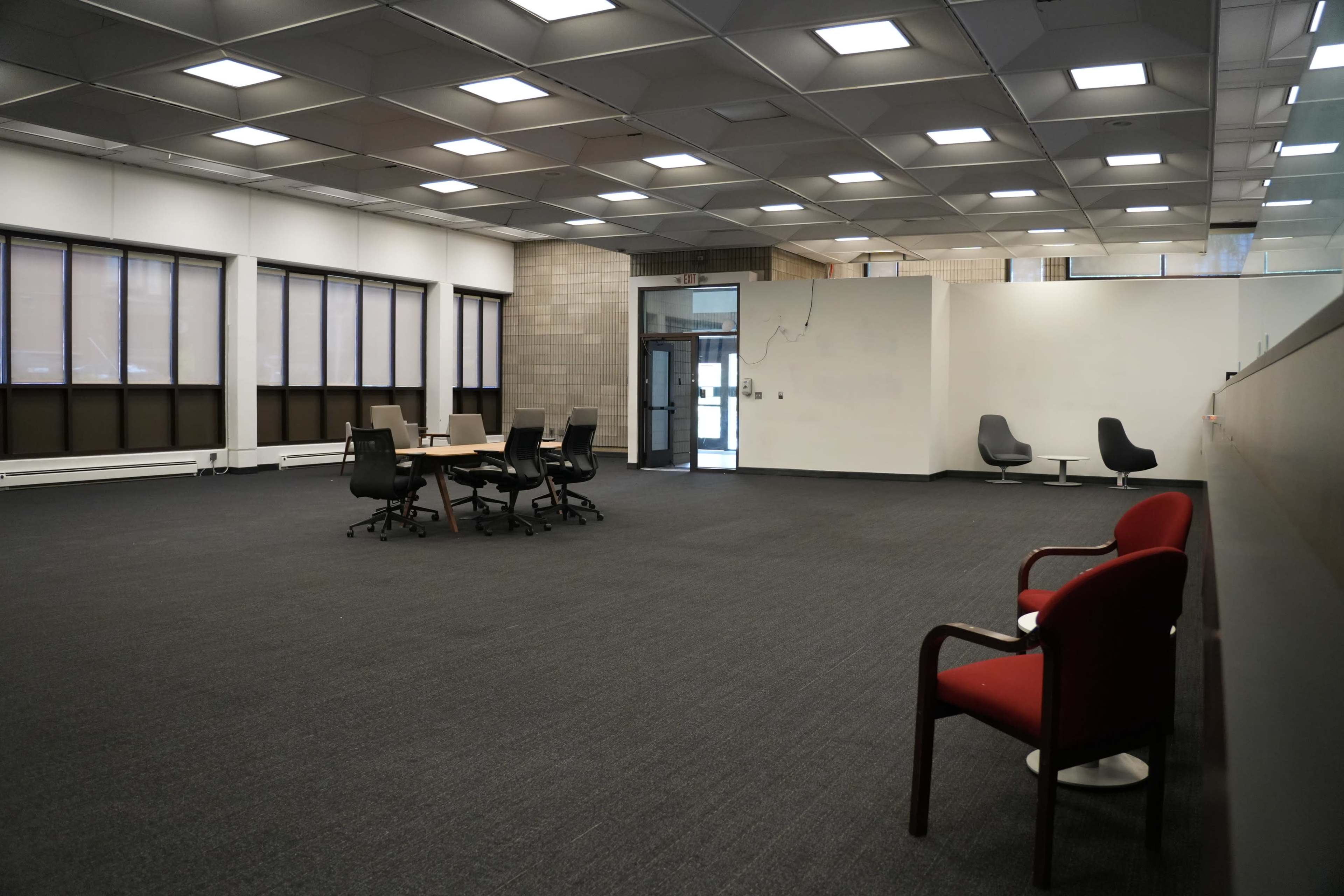 The image shows a spacious, empty room with a round table surrounded by chairs and two additional chairs positioned against a wall.