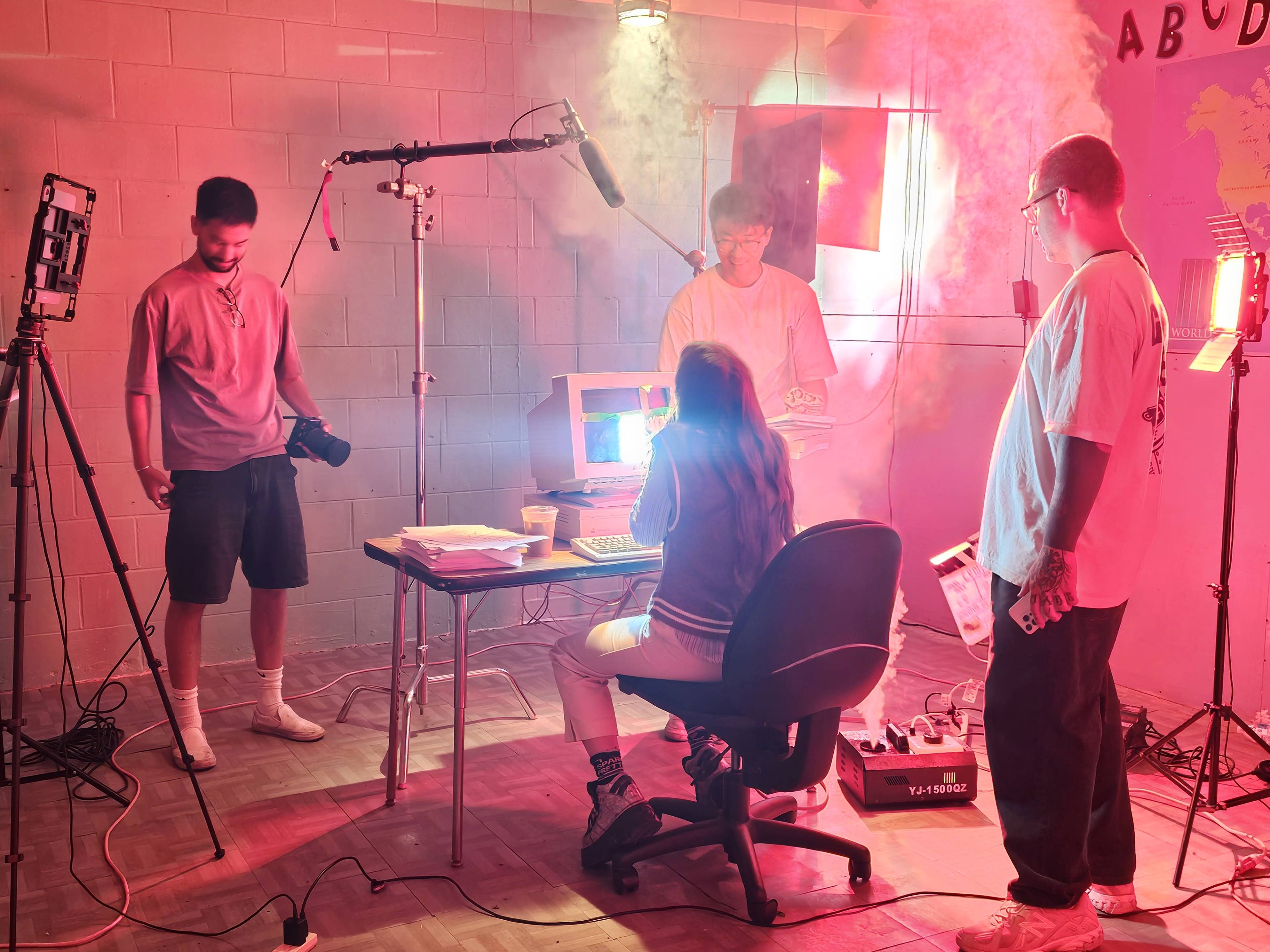 A group of four individuals is set up in a dimly lit room filled with smoke, with one person seated at a desk using an old computer while the others operate cameras and microphones.
