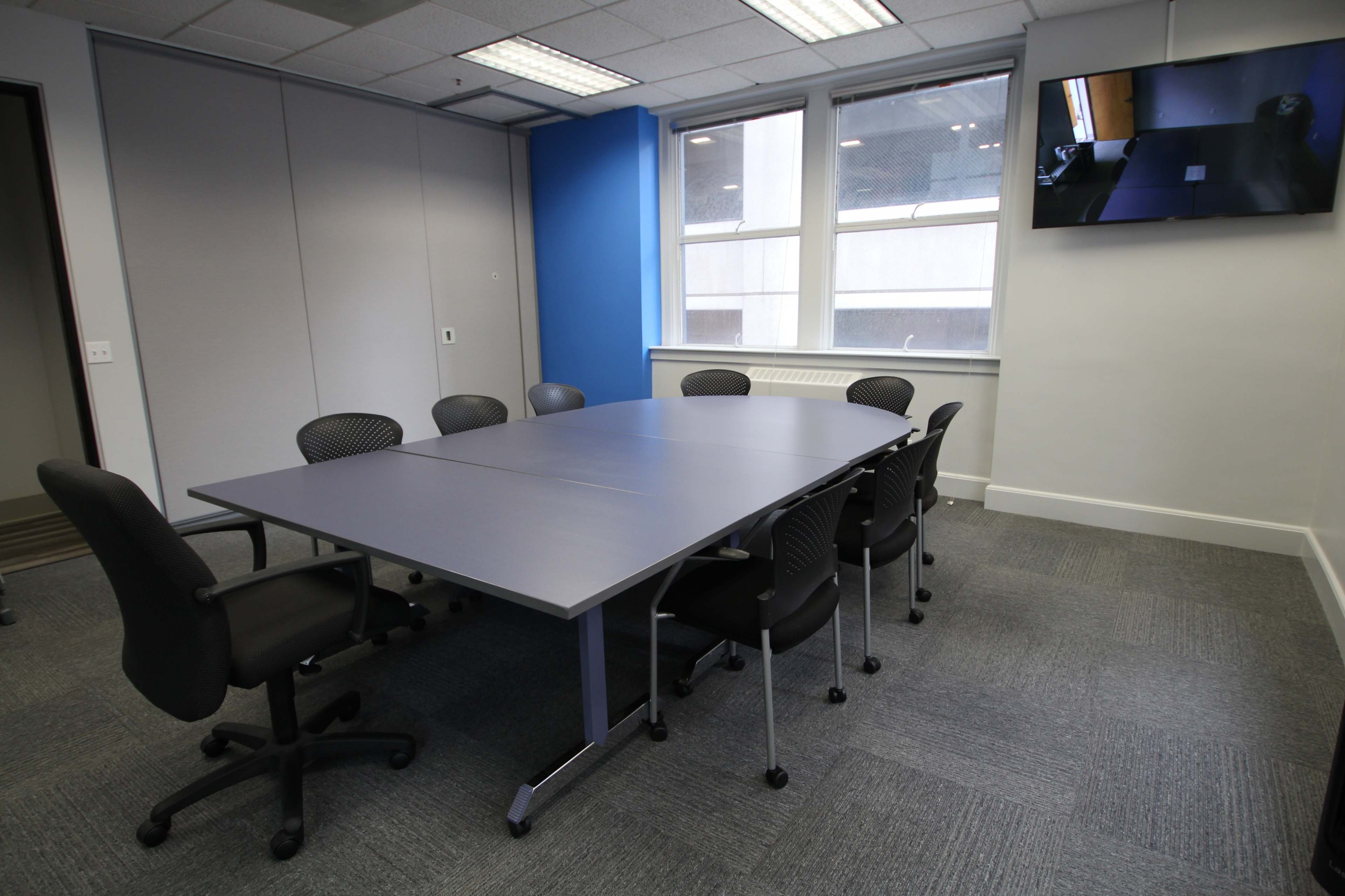 A meeting room features a long, rectangular table surrounded by black office chairs, with a mounted television screen on the wall.