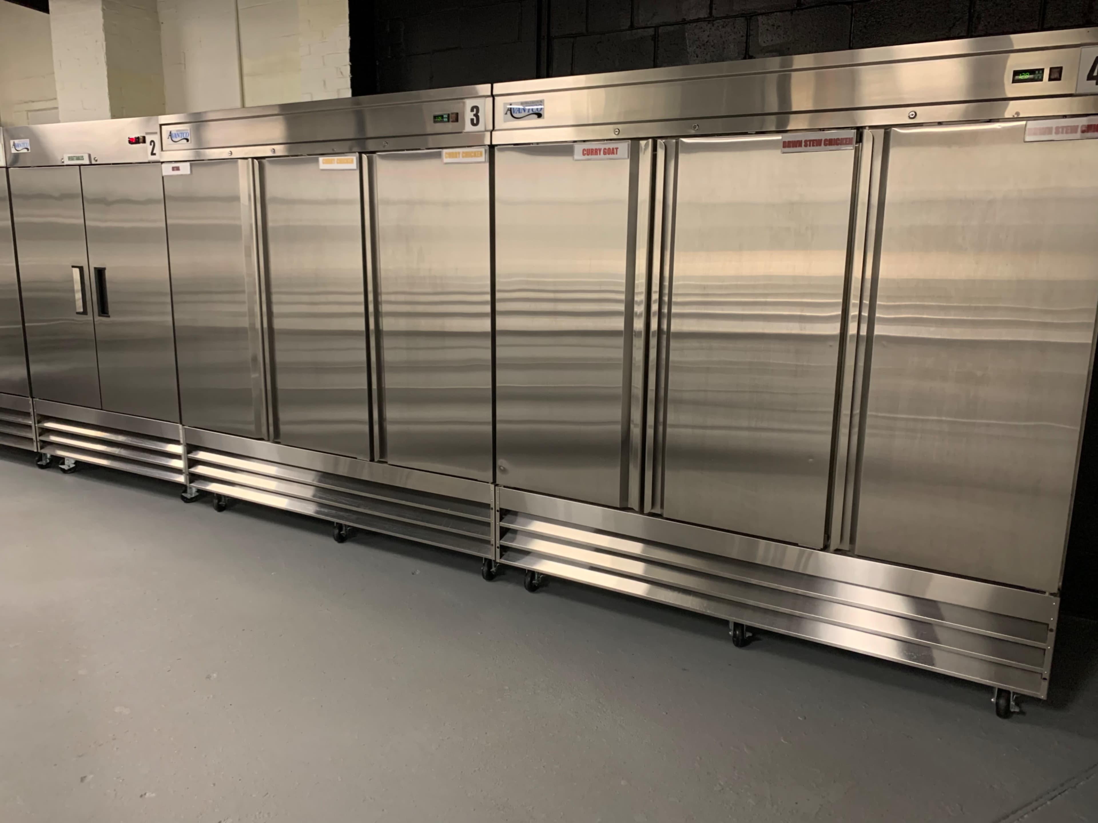 A row of several stainless steel commercial refrigerators with glass doors in a dimly lit room.