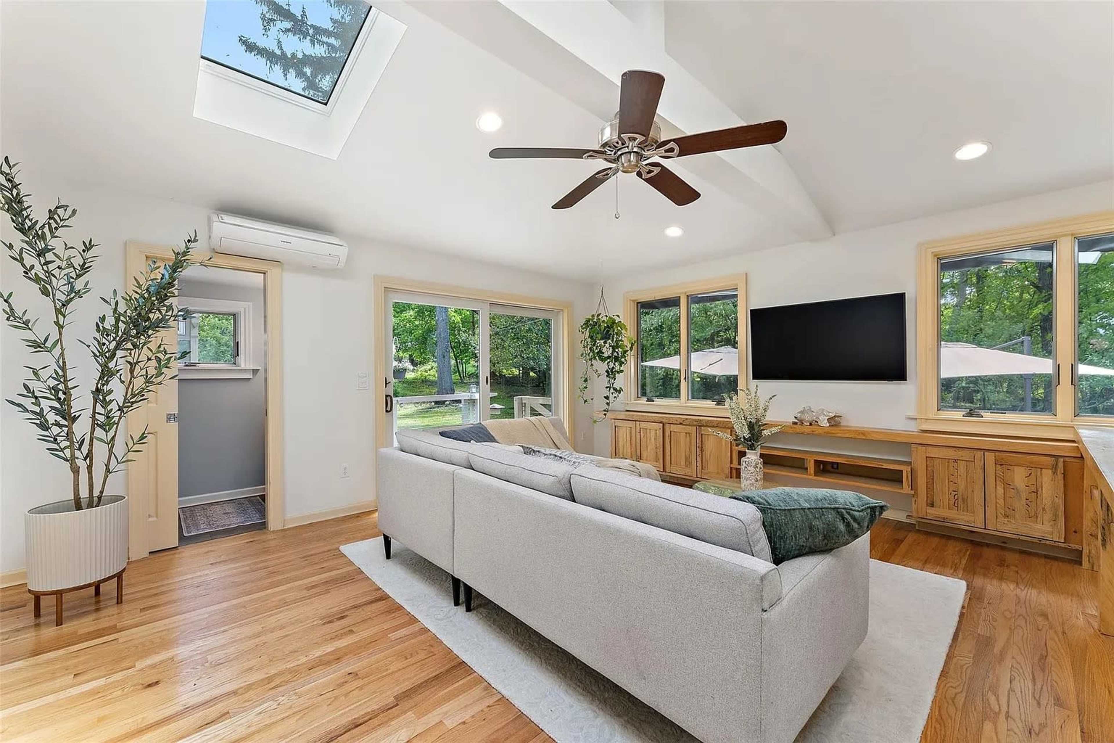 The living room features a light-colored sofa, large windows with a view of greenery, and a ceiling fan, with a wooden entertainment unit and a skylight above.