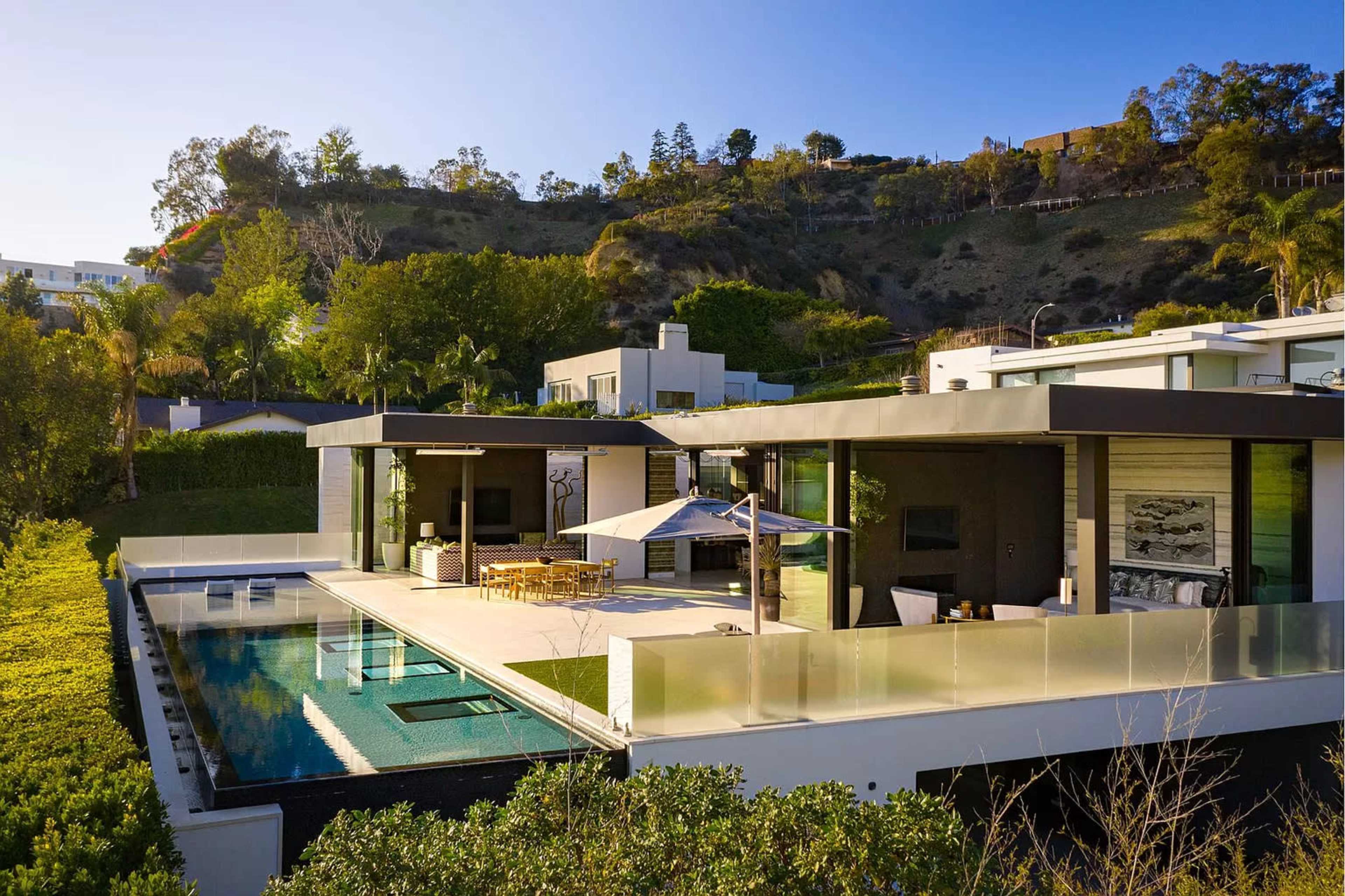 The image shows a modern, two-story house overlooking a pool area, surrounded by lush landscaping and hills.