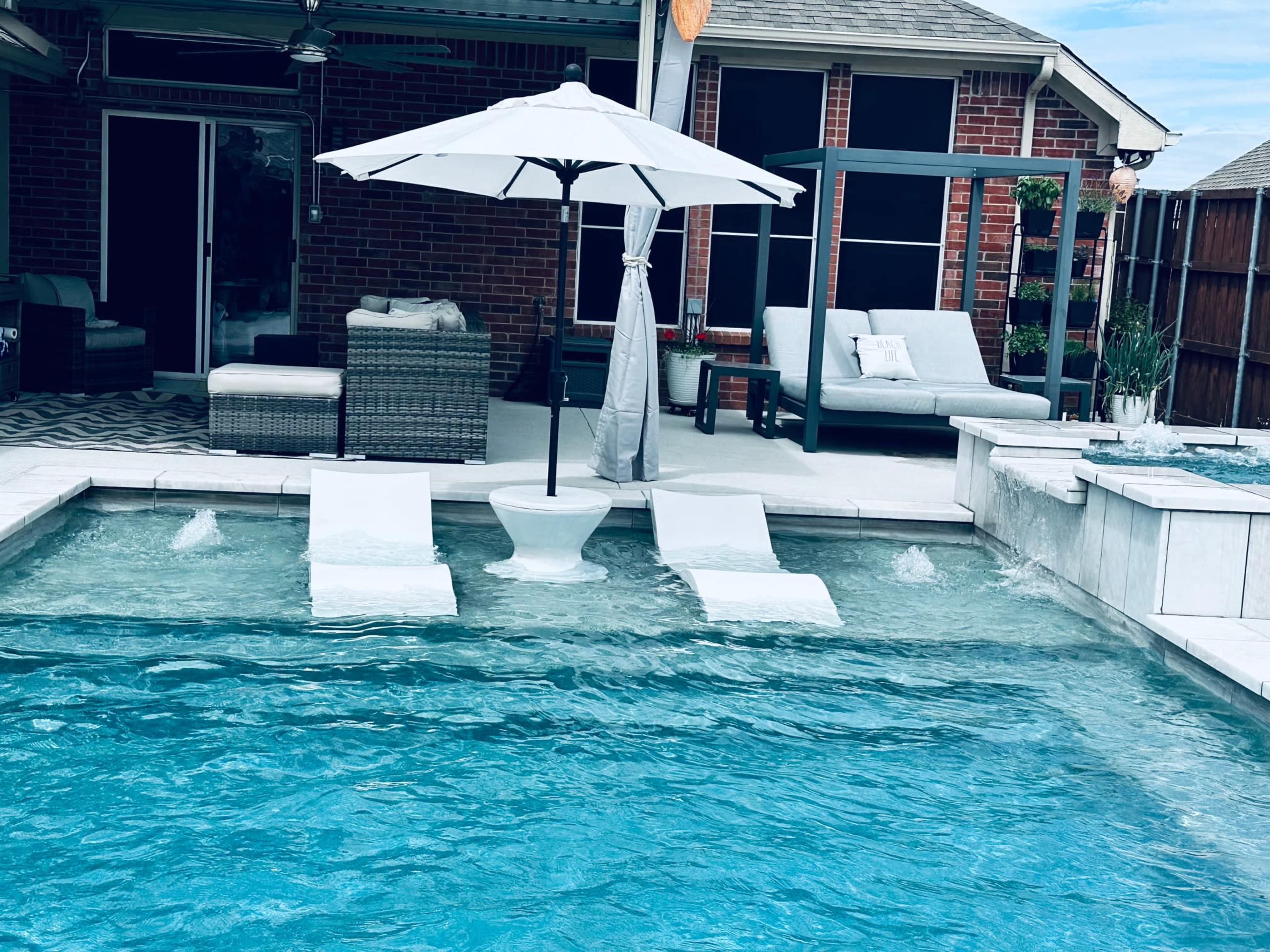 The image shows a backyard pool area featuring two white lounge chairs partially submerged in the water beneath a large umbrella, with a brick patio and seating arrangement in the background.