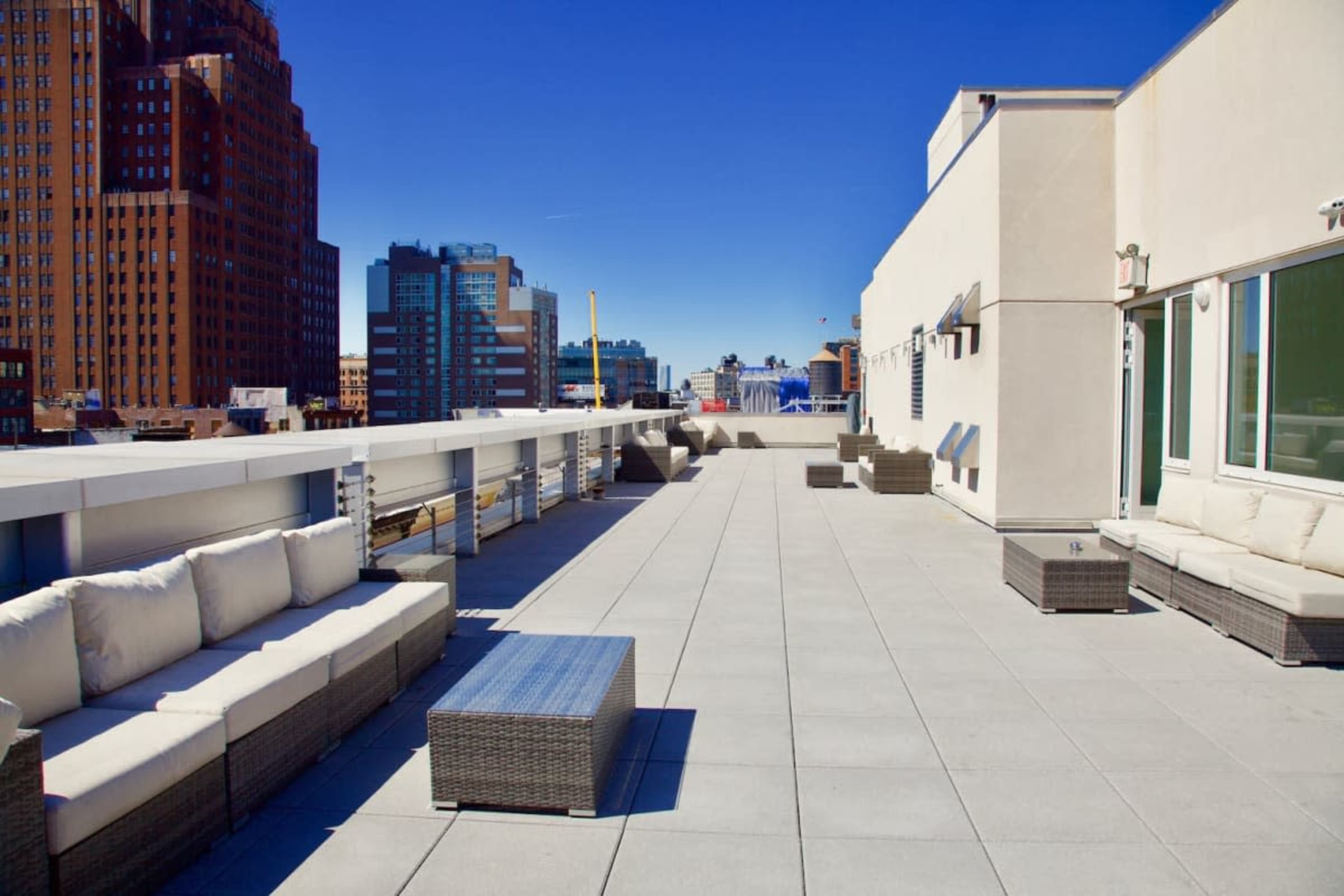 The image shows a spacious rooftop terrace with white sofas and glass tables overlooking a city skyline under a clear blue sky.