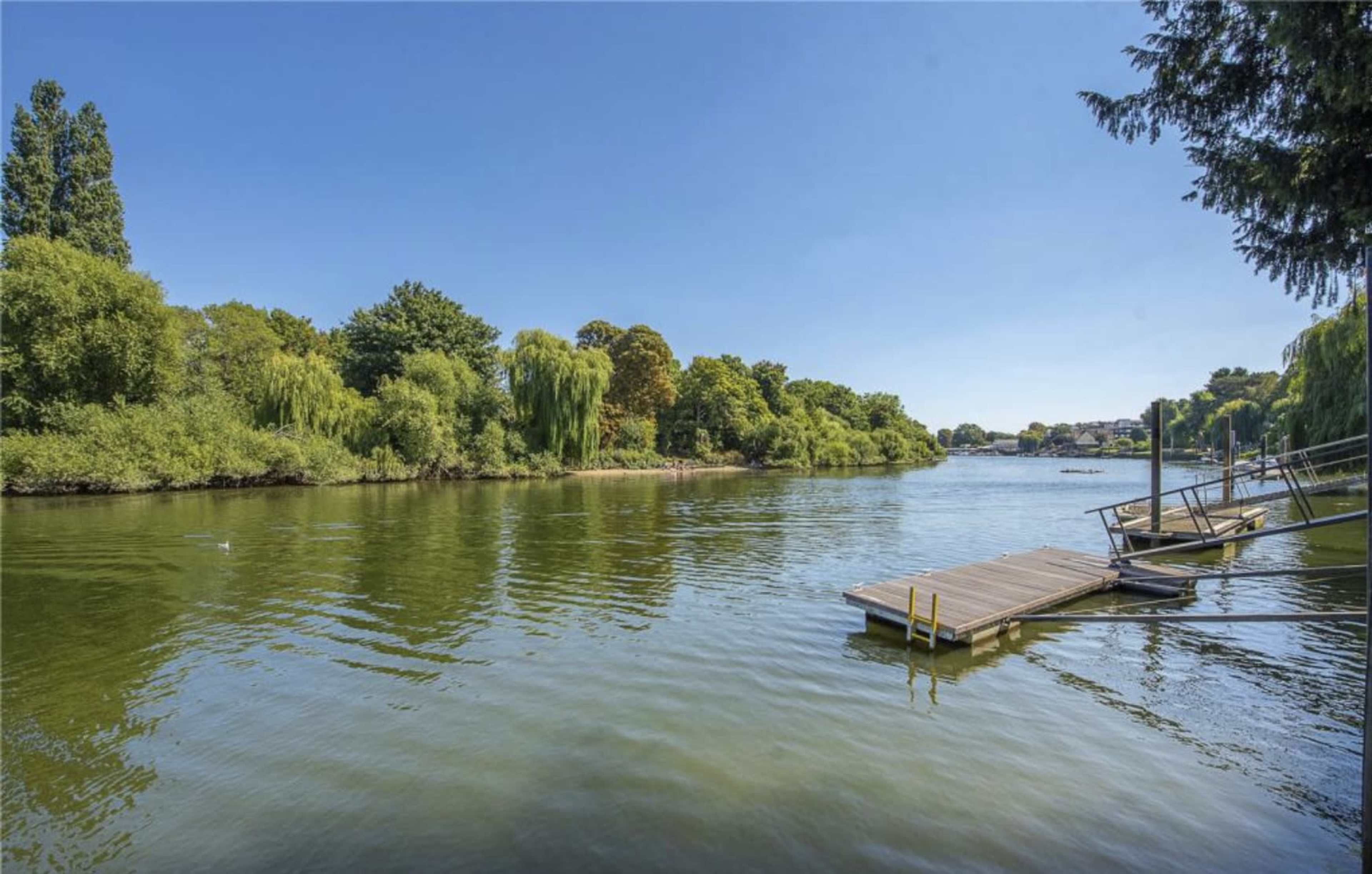 A wooden dock extends into a calm river surrounded by lush greenery under a clear blue sky.