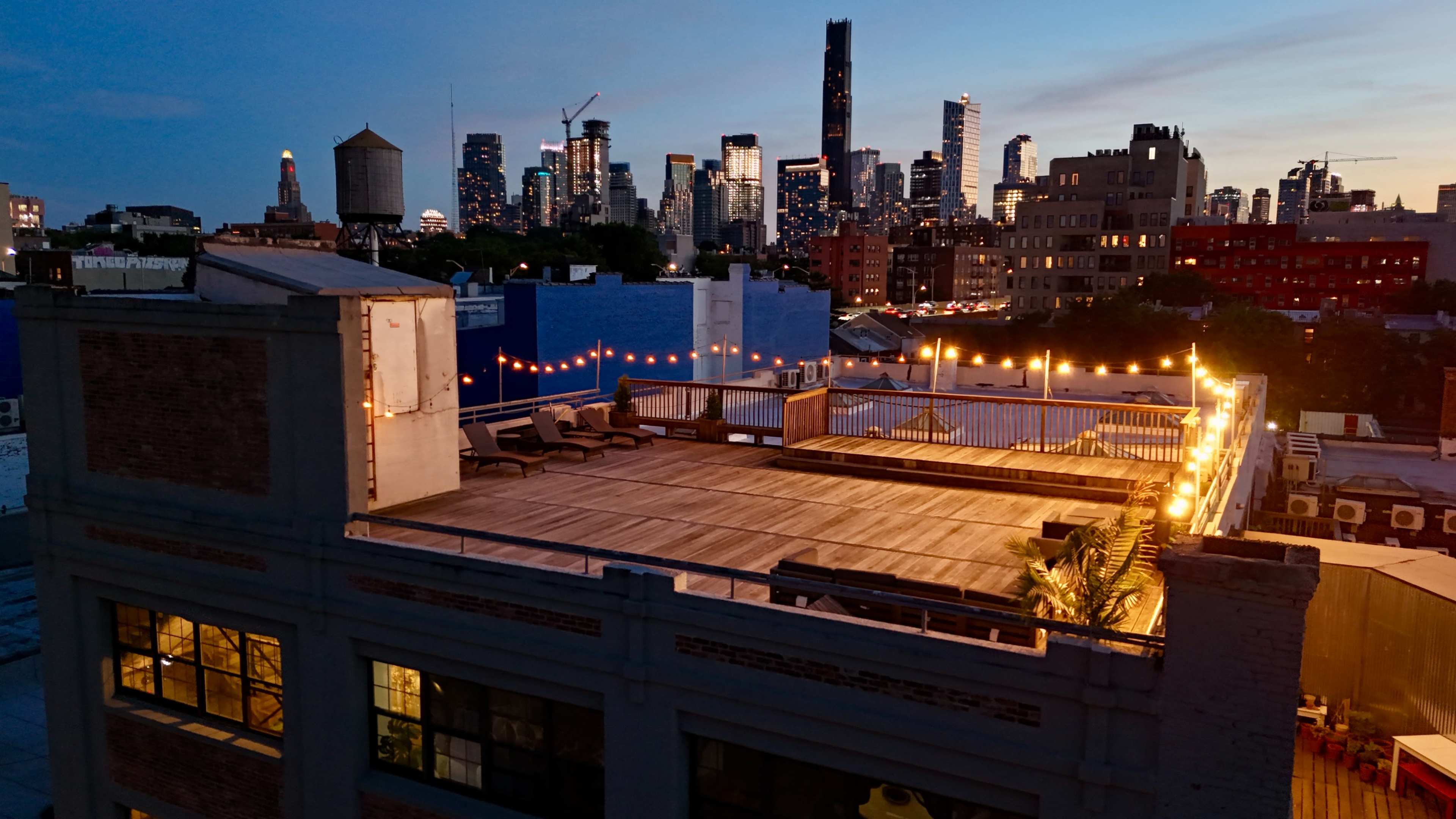 A rooftop space with a wooden deck and string lights, overlooking a skyline of tall buildings at dusk.