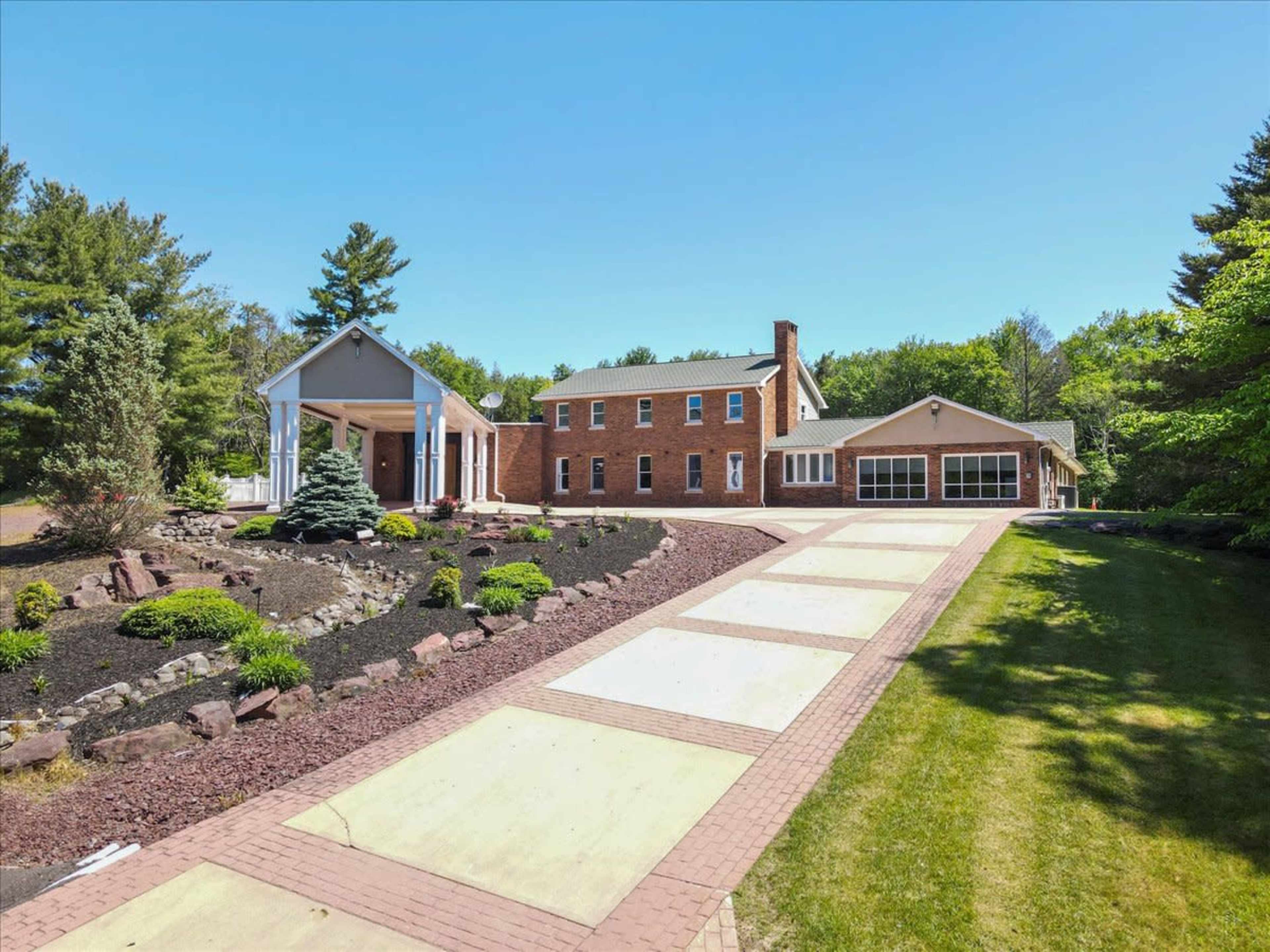 A large brick house with a white columned entrance, surrounded by landscaped gardens and a paved driveway leading to a garage.