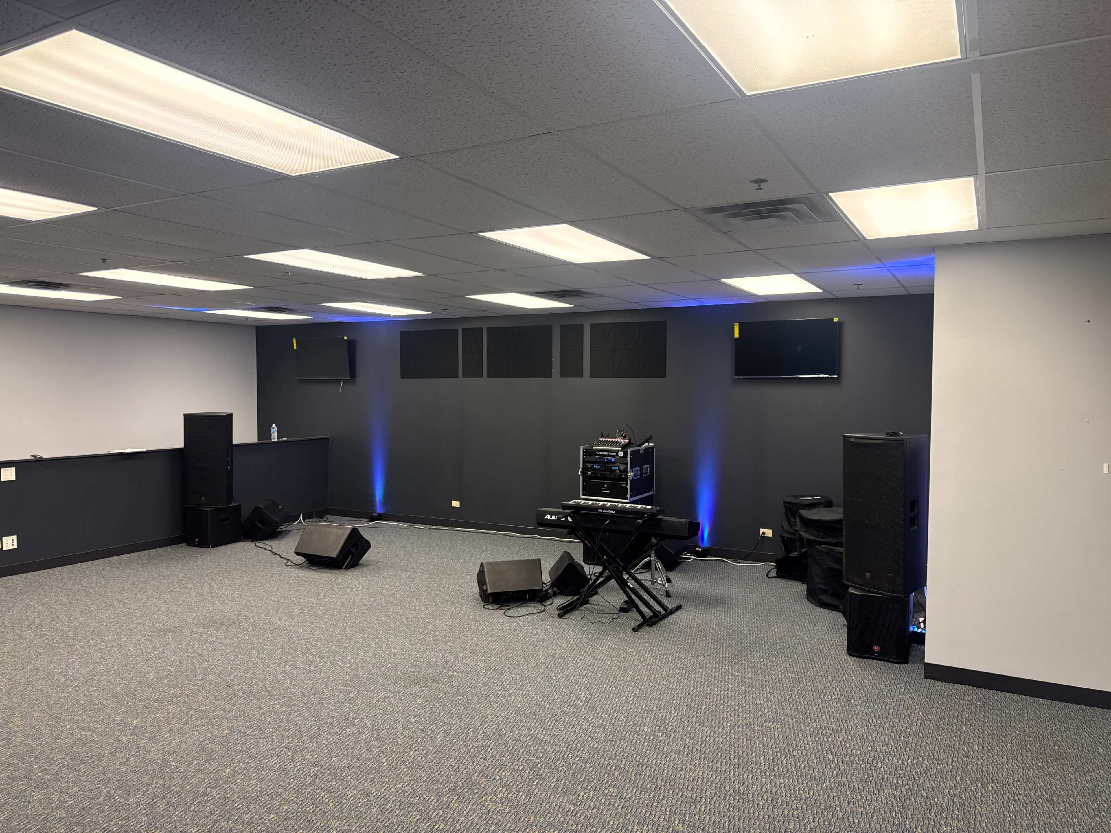 The image shows a performance space with a stage setup that includes speakers, a keyboard, and two wall-mounted screens.