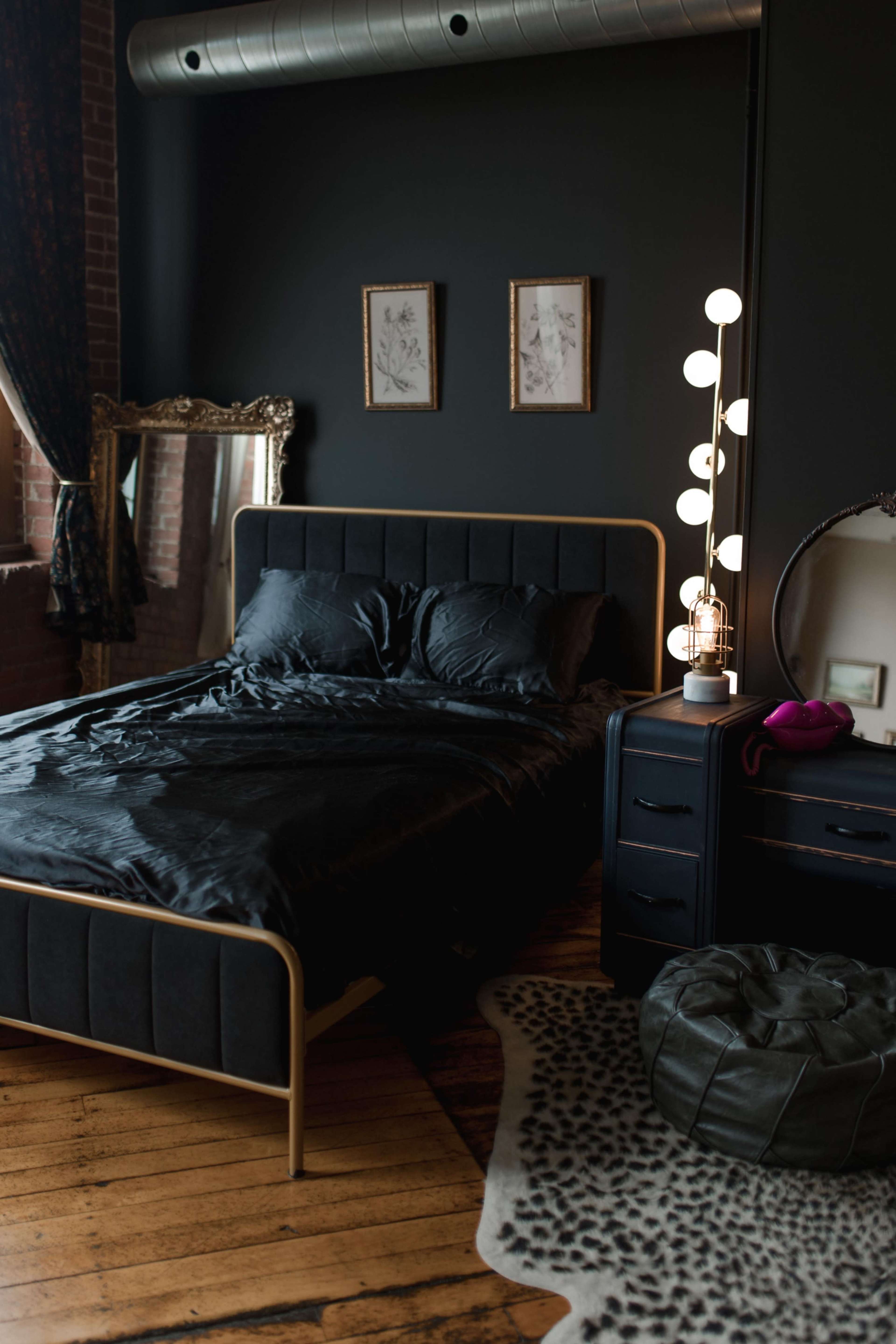 The image shows a modern bedroom with a black bedspread, a large mirror, a decorative floor lamp, and a light-colored animal print rug.