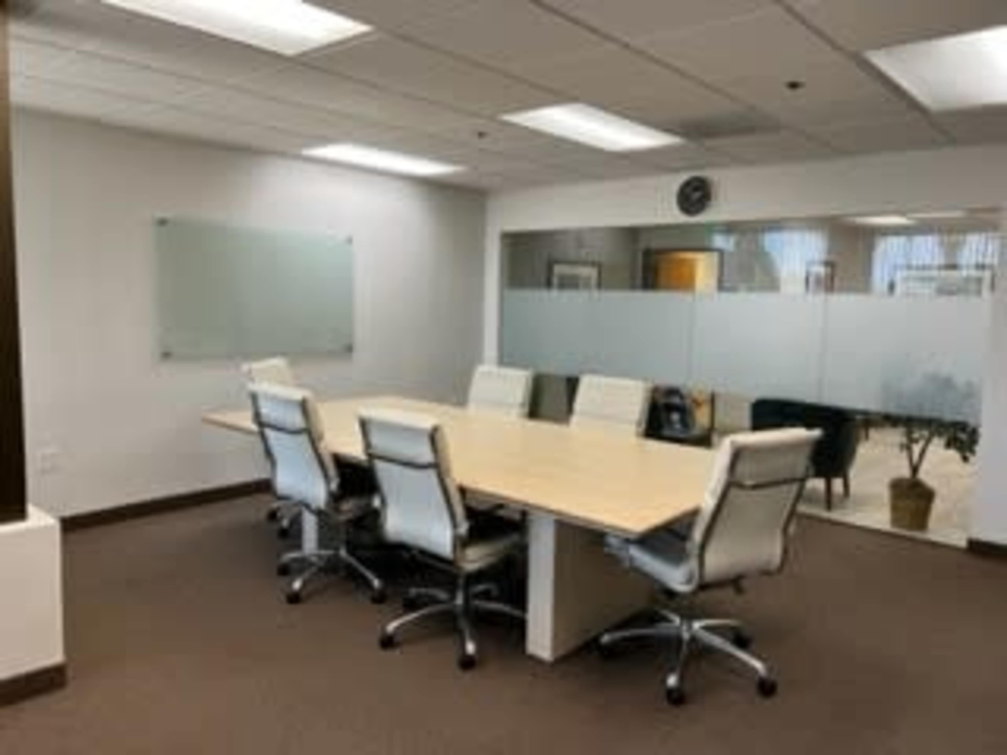 A spacious conference room features a long wooden table surrounded by six white office chairs, with glass partitions and a plant visible in the background.