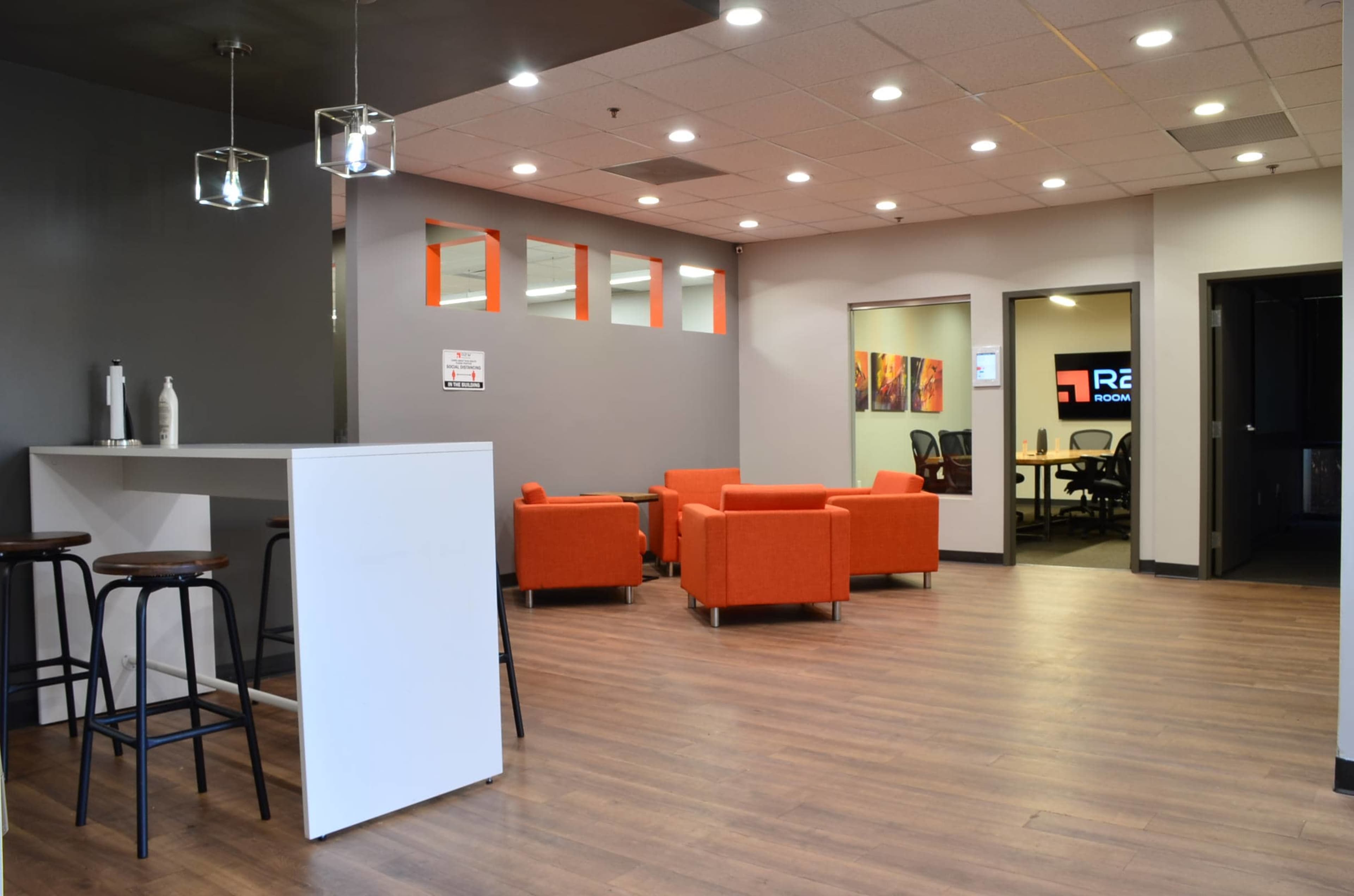 A modern office interior with orange chairs, a white counter, and a meeting room visible in the background.