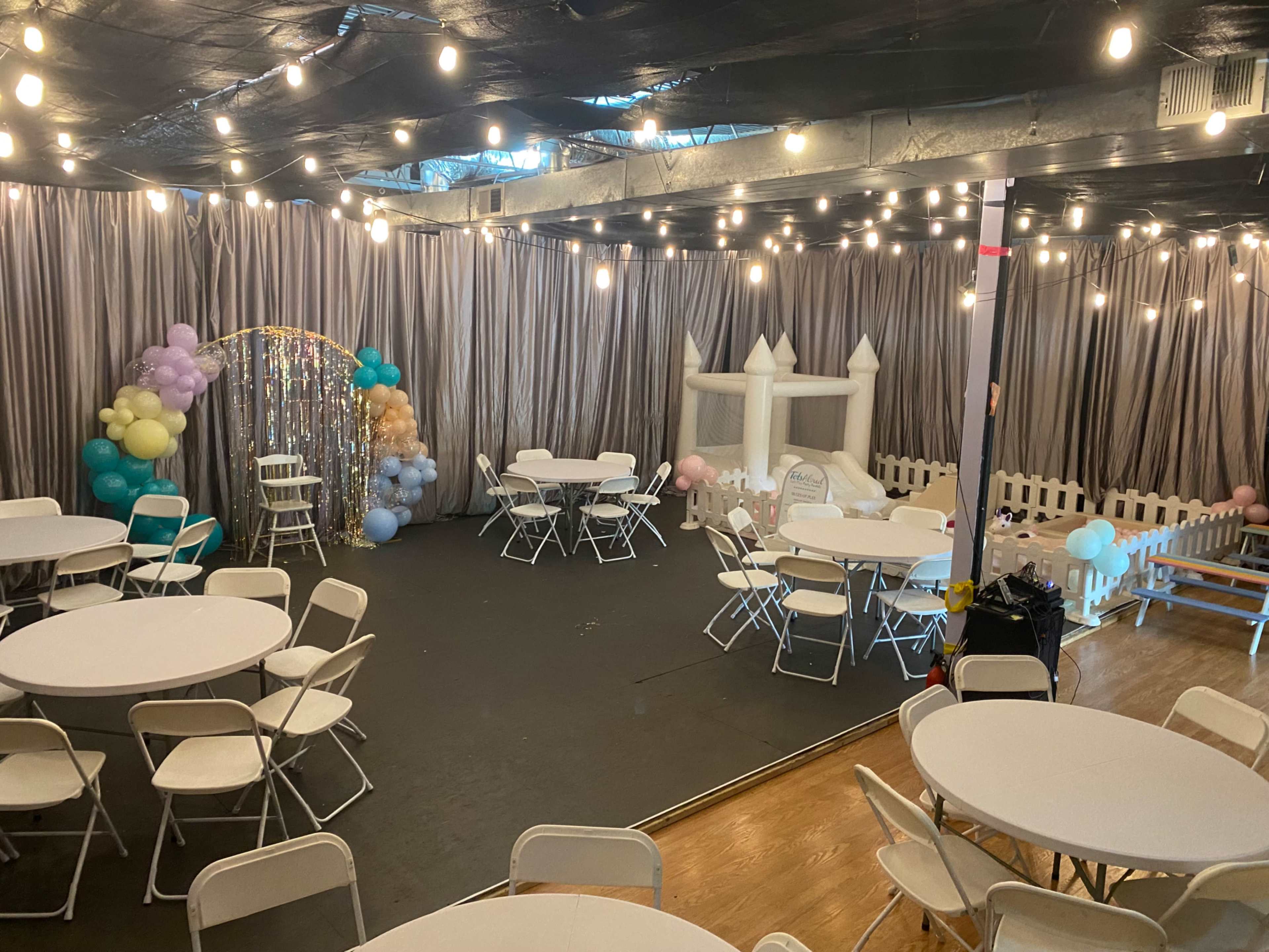 A decorated event space features circular tables, chairs, colorful balloon arrangements, and a bounce house.