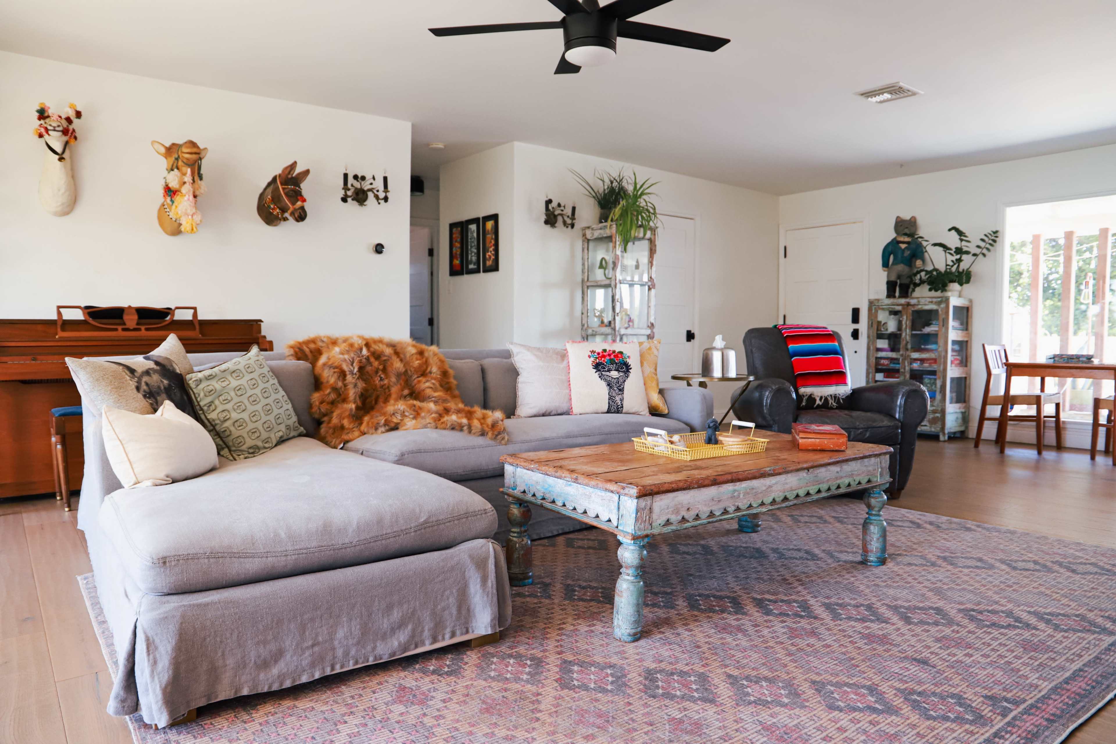 The living room features a gray sectional sofa, a wooden coffee table with decorative items, and a piano against the wall, with various art pieces and plants in the space.