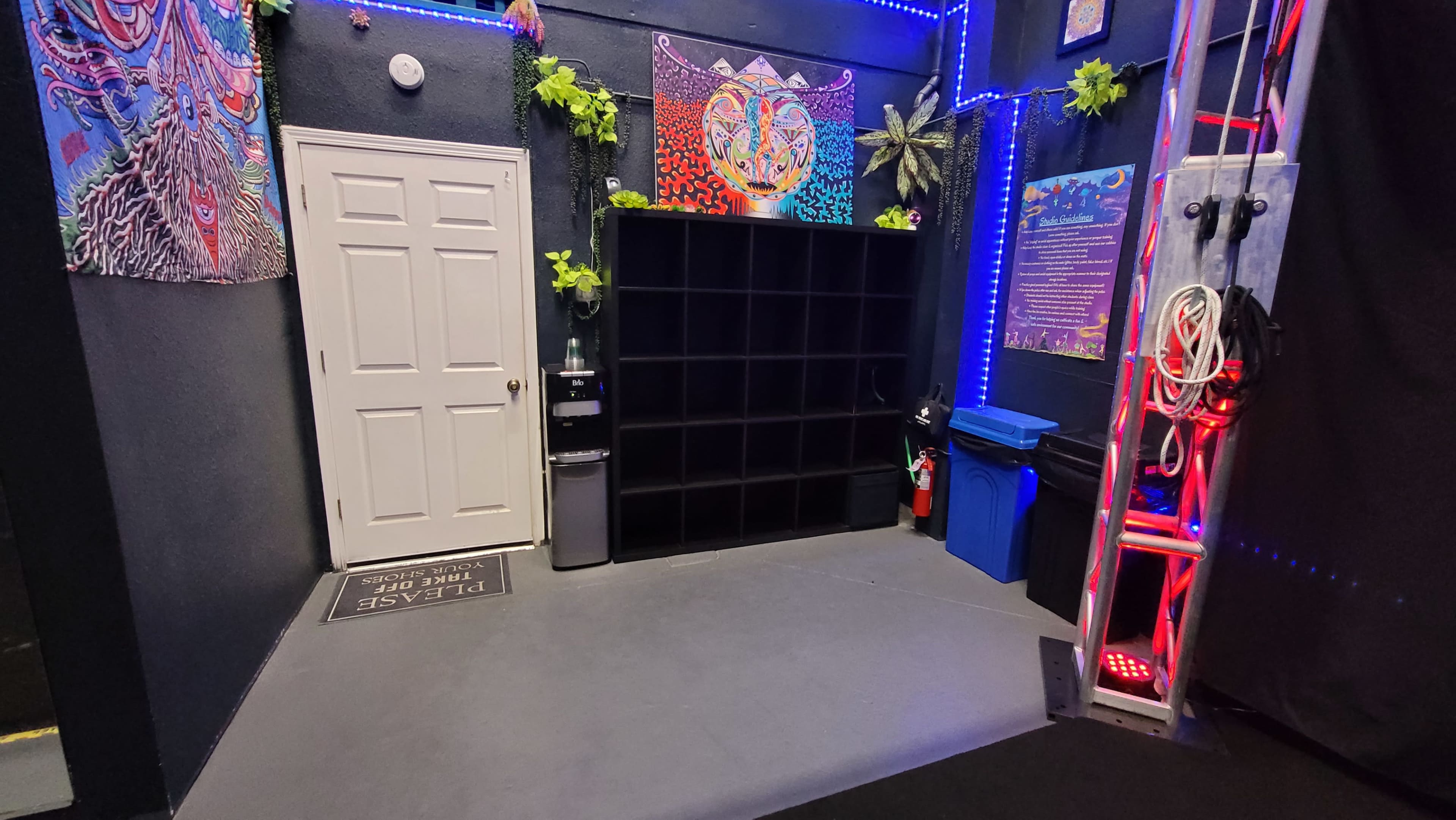 A small room featuring a white door, a black cubical storage unit, a water cooler, a blue recycling bin, and colorful wall decorations illuminated by blue lights.