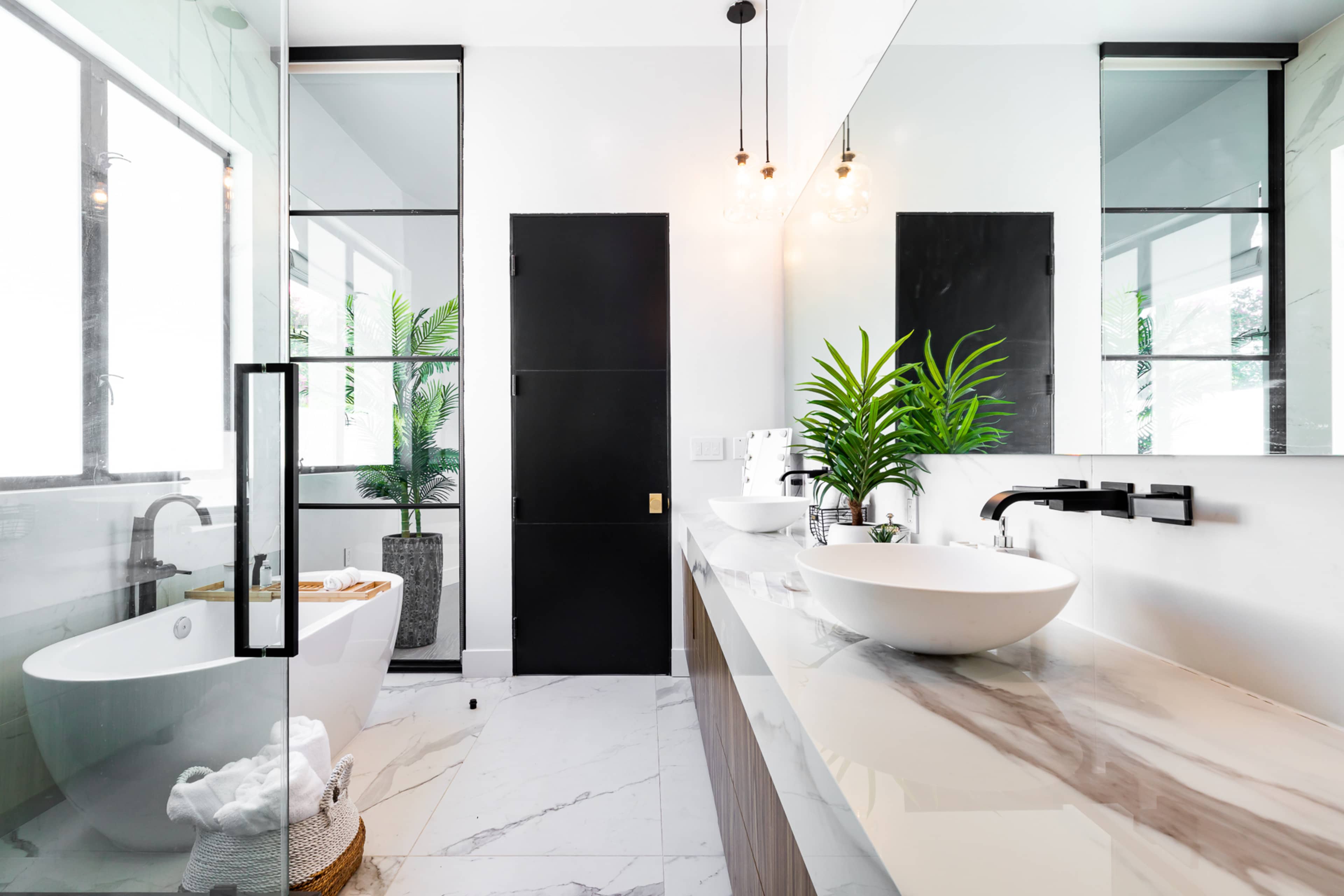 The image shows a modern bathroom featuring a freestanding bathtub, a large sink on a marble countertop, glass shower doors, and potted plants for decoration.