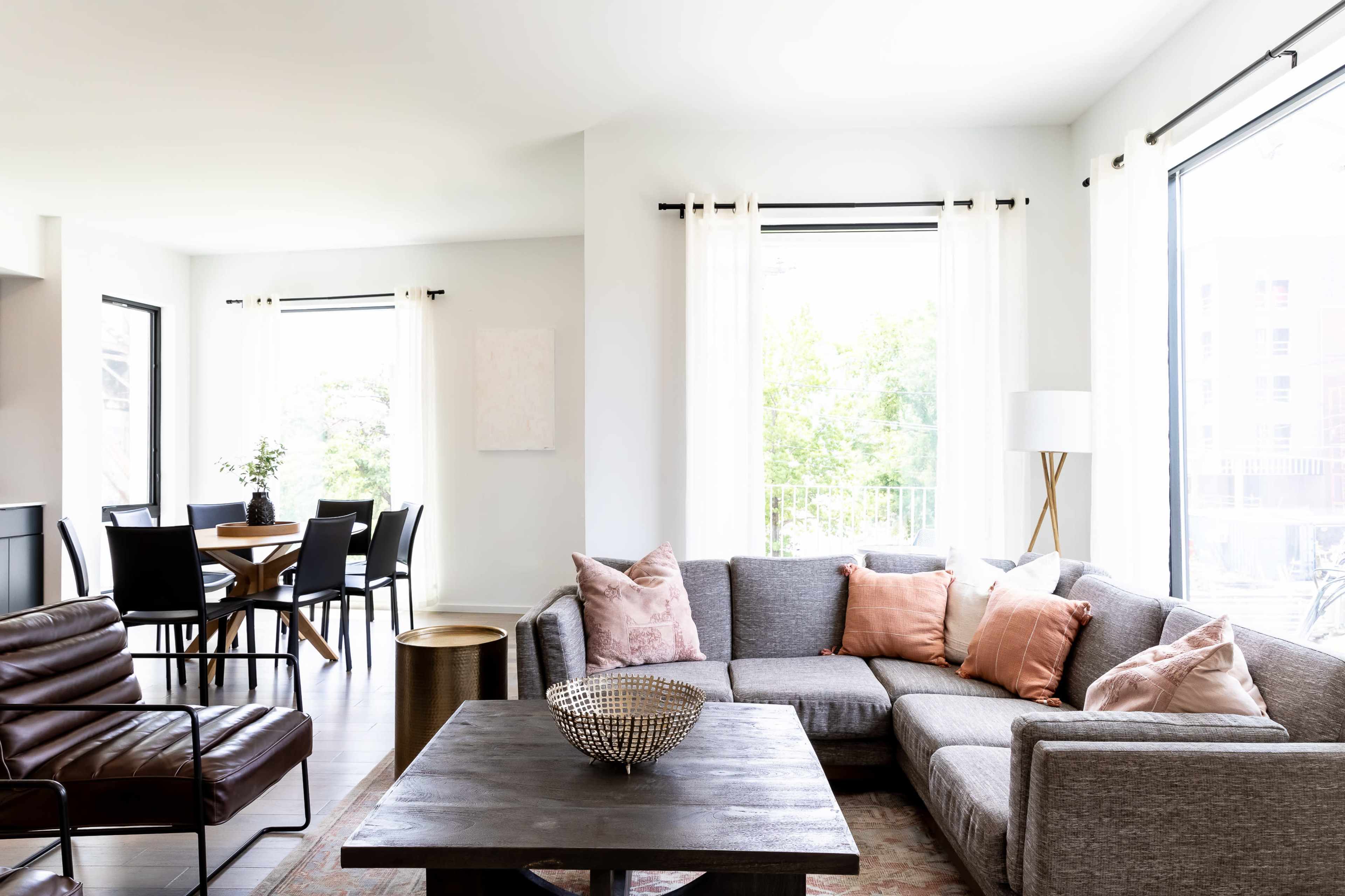 A bright living room with a gray sectional sofa, a dark wooden coffee table, and a dining area featuring a round table and black chairs.