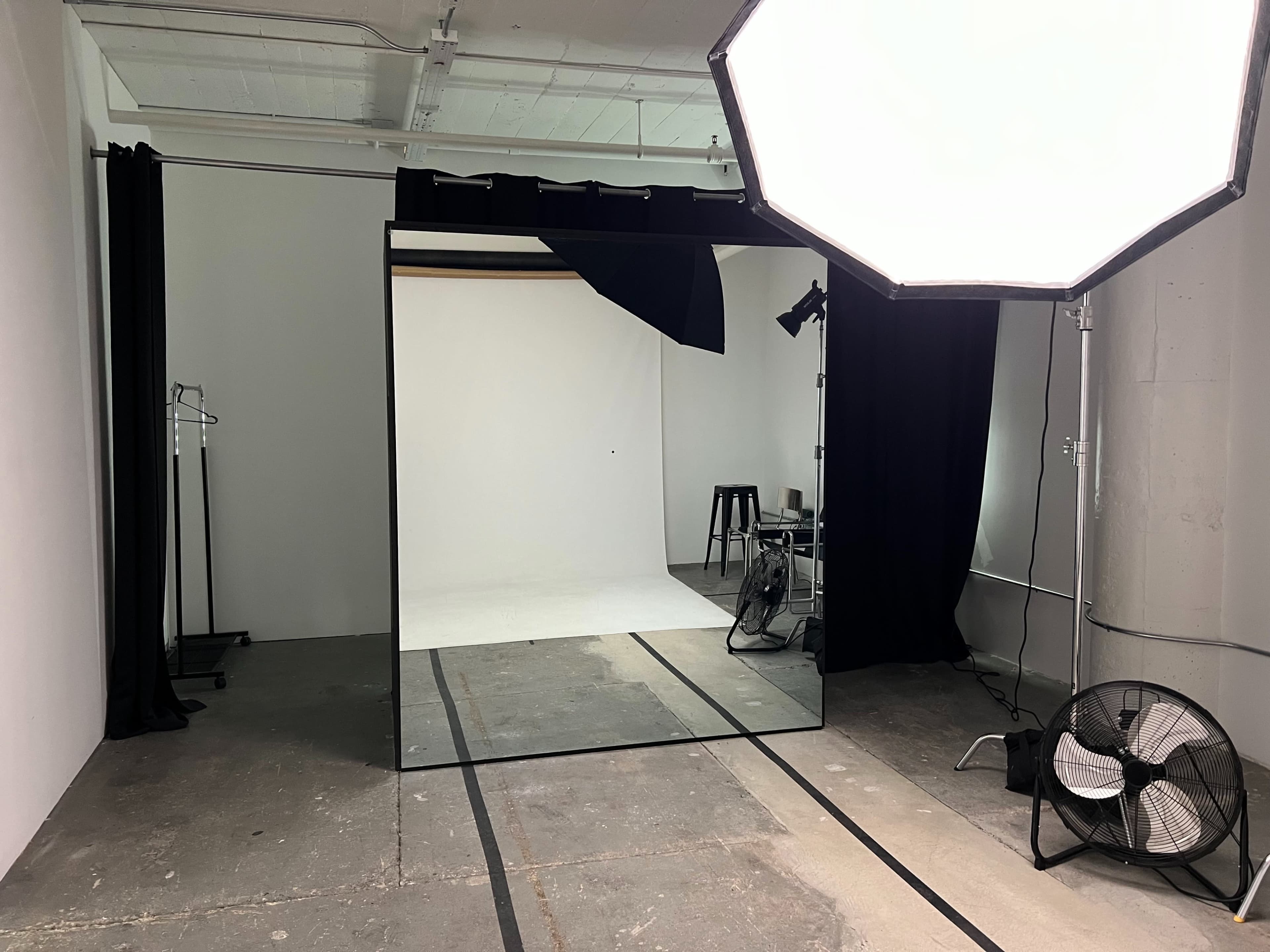 The image shows a photography studio space with a large mirror, backdrop, lighting equipment, and a fan.
