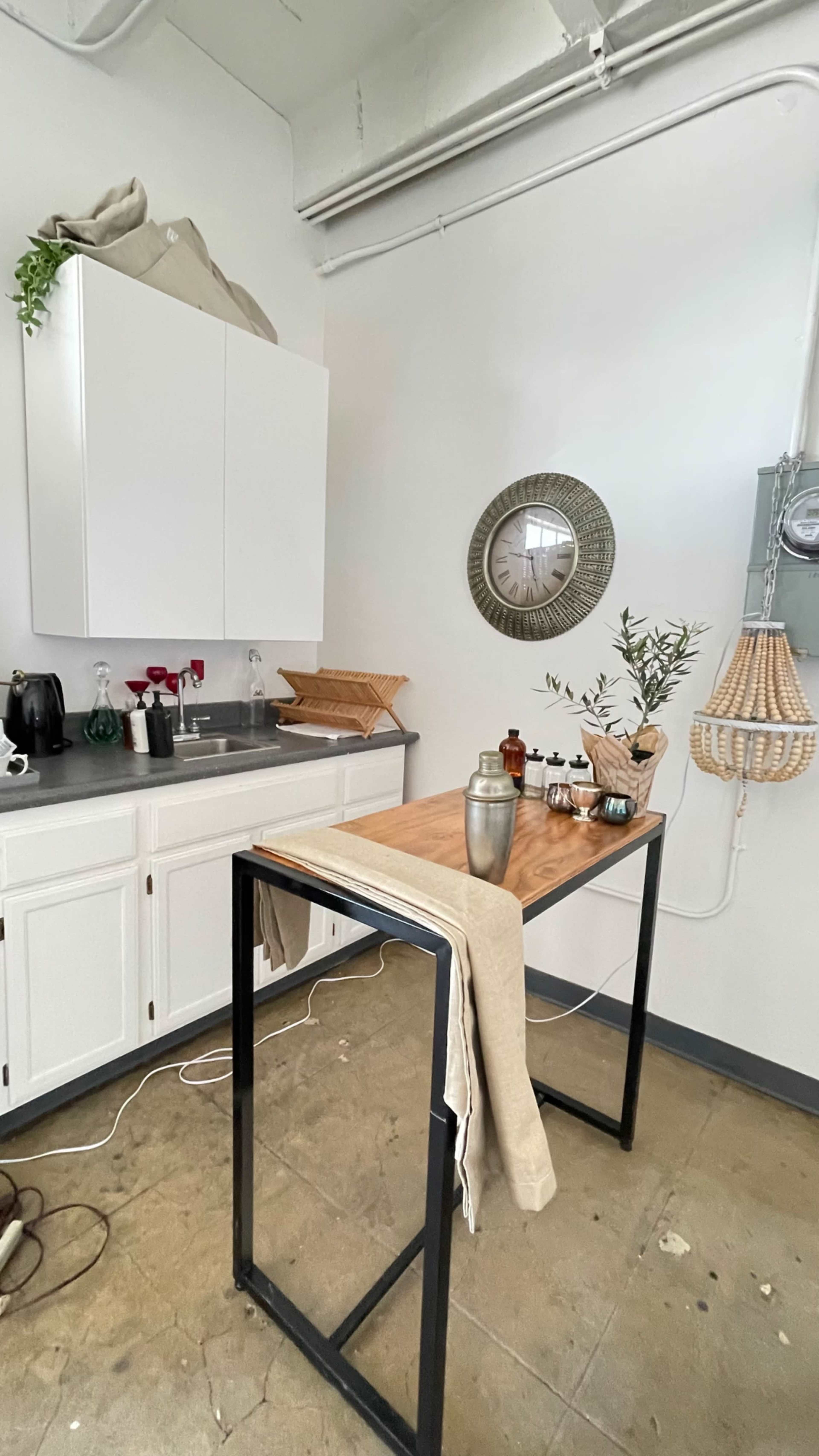 A minimalist kitchen area features a wooden table, a wall clock, a sink, and various kitchen items on shelves.