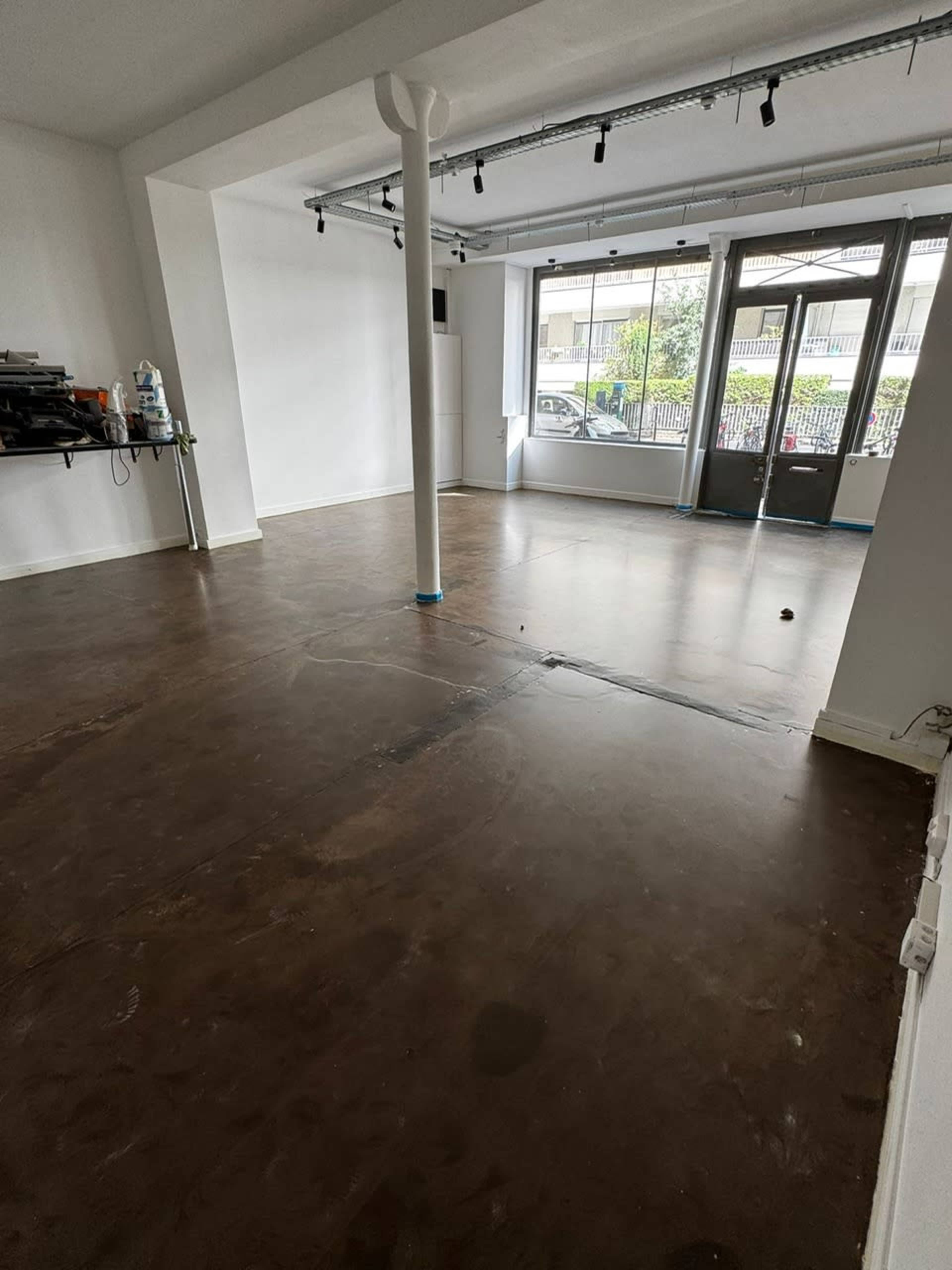 The image shows a spacious, empty room with a polished concrete floor, a large window, and bright overhead lights.