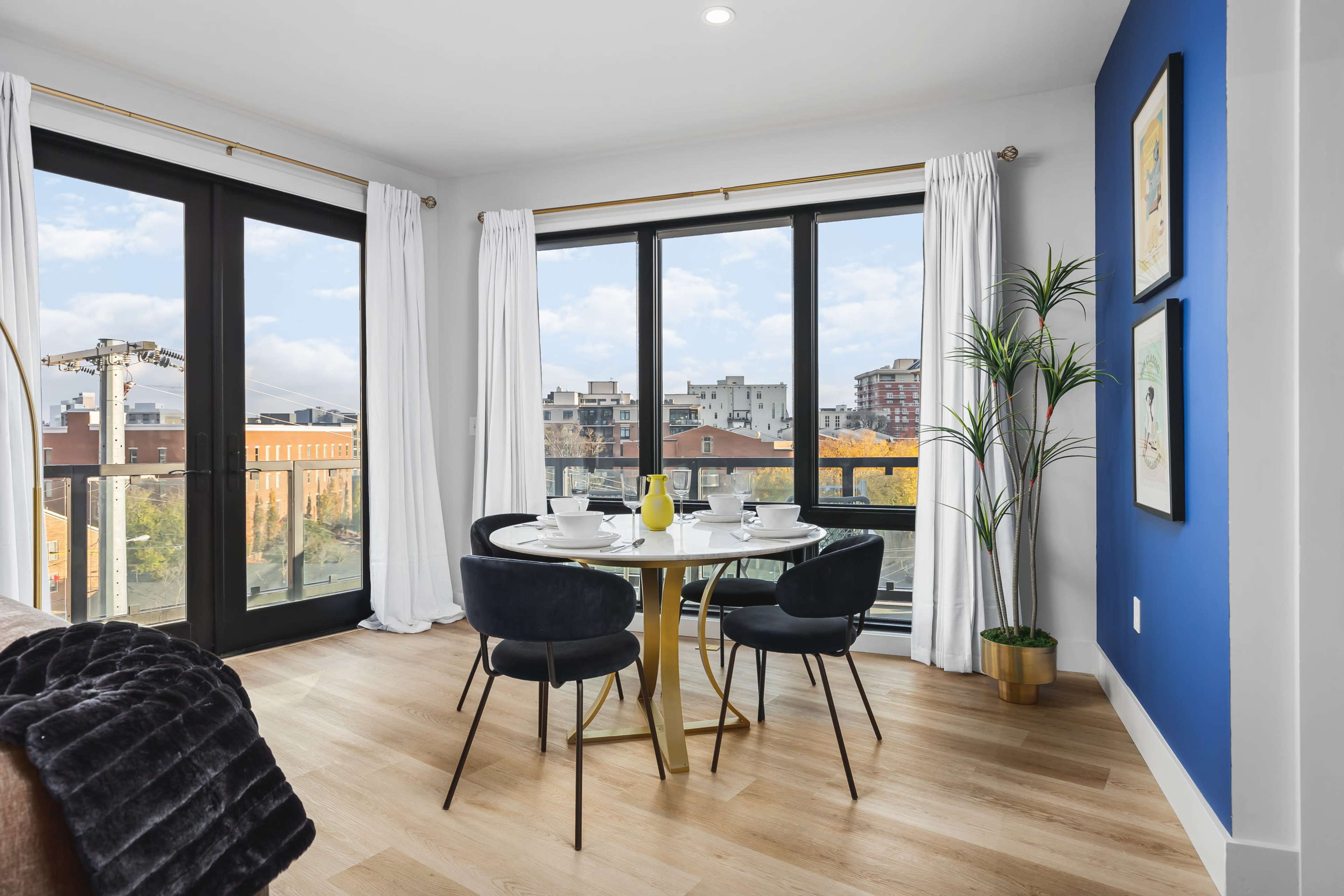 A dining area features a round table set for four, with large windows showcasing an urban view and light-colored curtains.