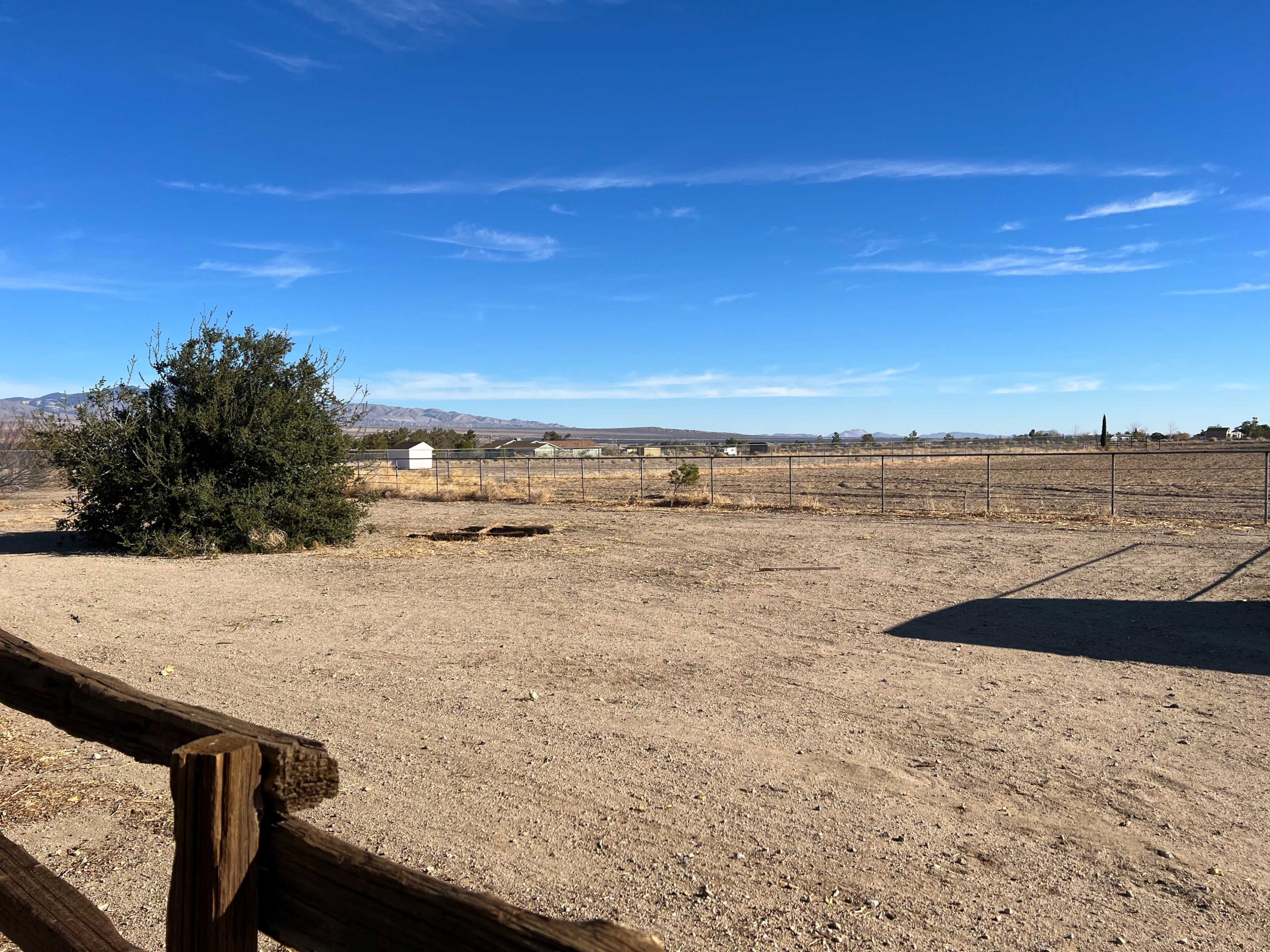 A wide, open landscape shows a barren area with sparse vegetation and a fence surrounding distant structures against a clear sky.