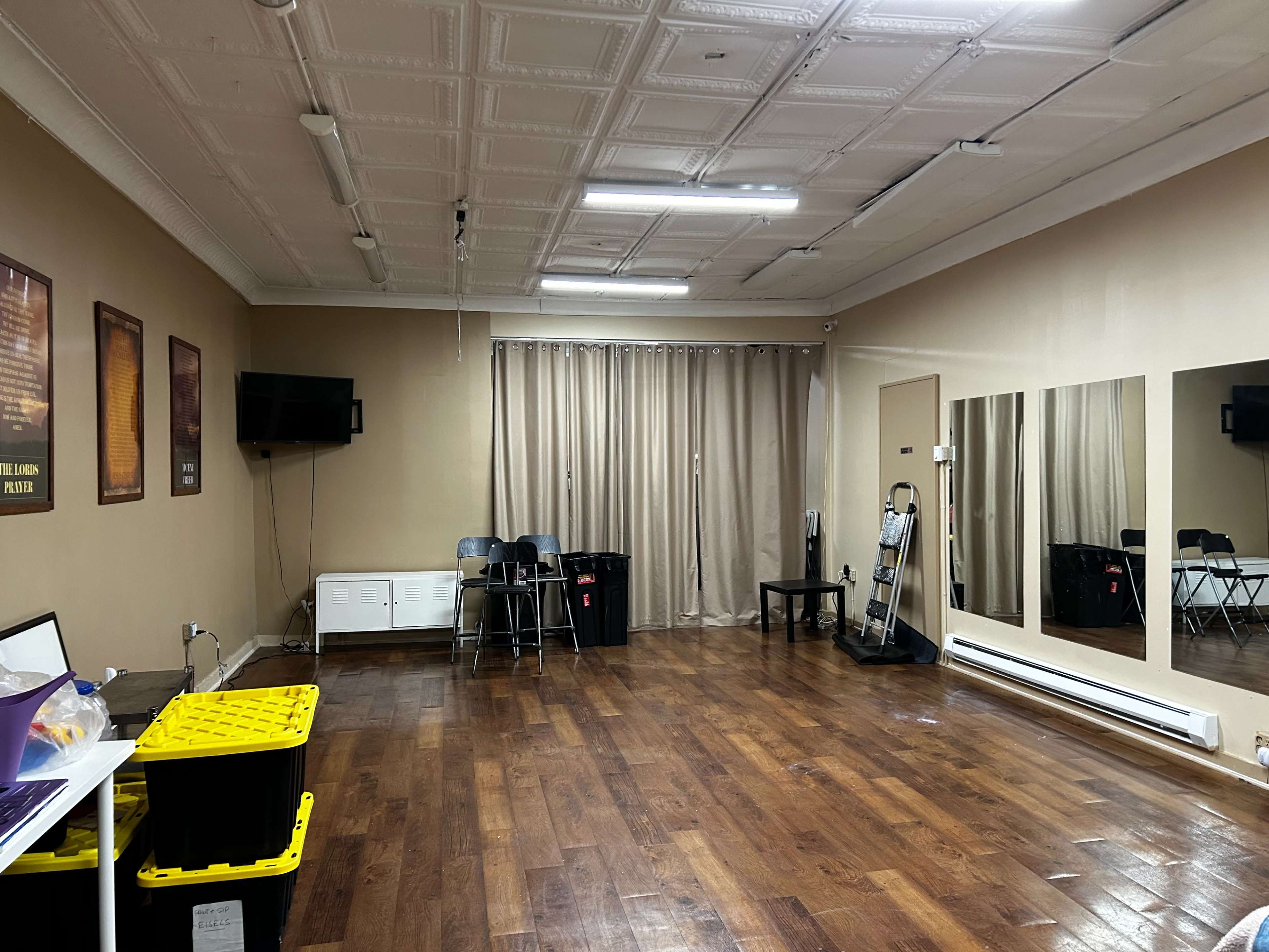 A spacious room with wooden flooring, a large mirror on one wall, a television mounted on another, and several chairs and storage boxes arranged around the space.
