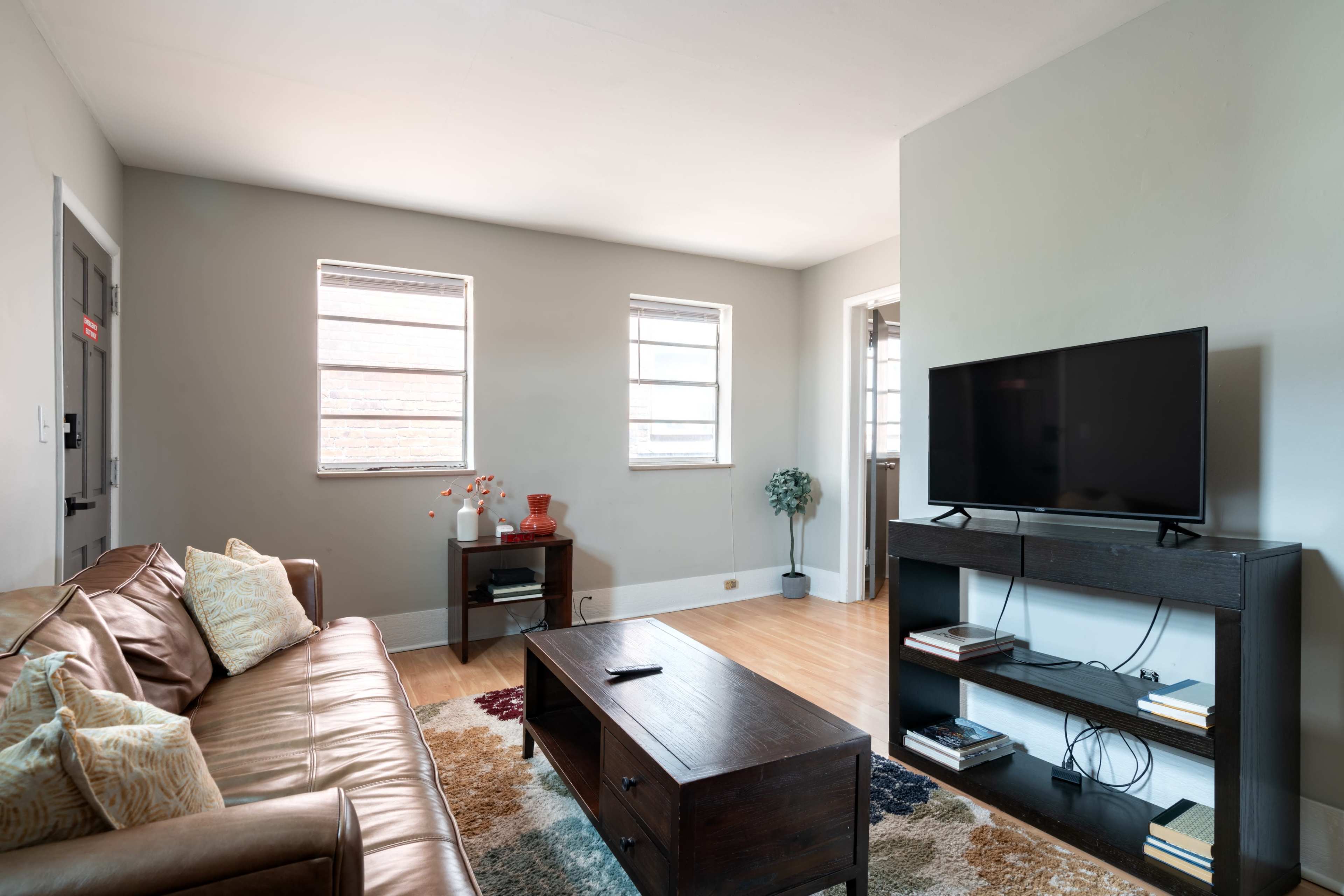 The room features a brown leather sofa, a wooden coffee table, and a black television on a shelf, with windows allowing natural light to enter.