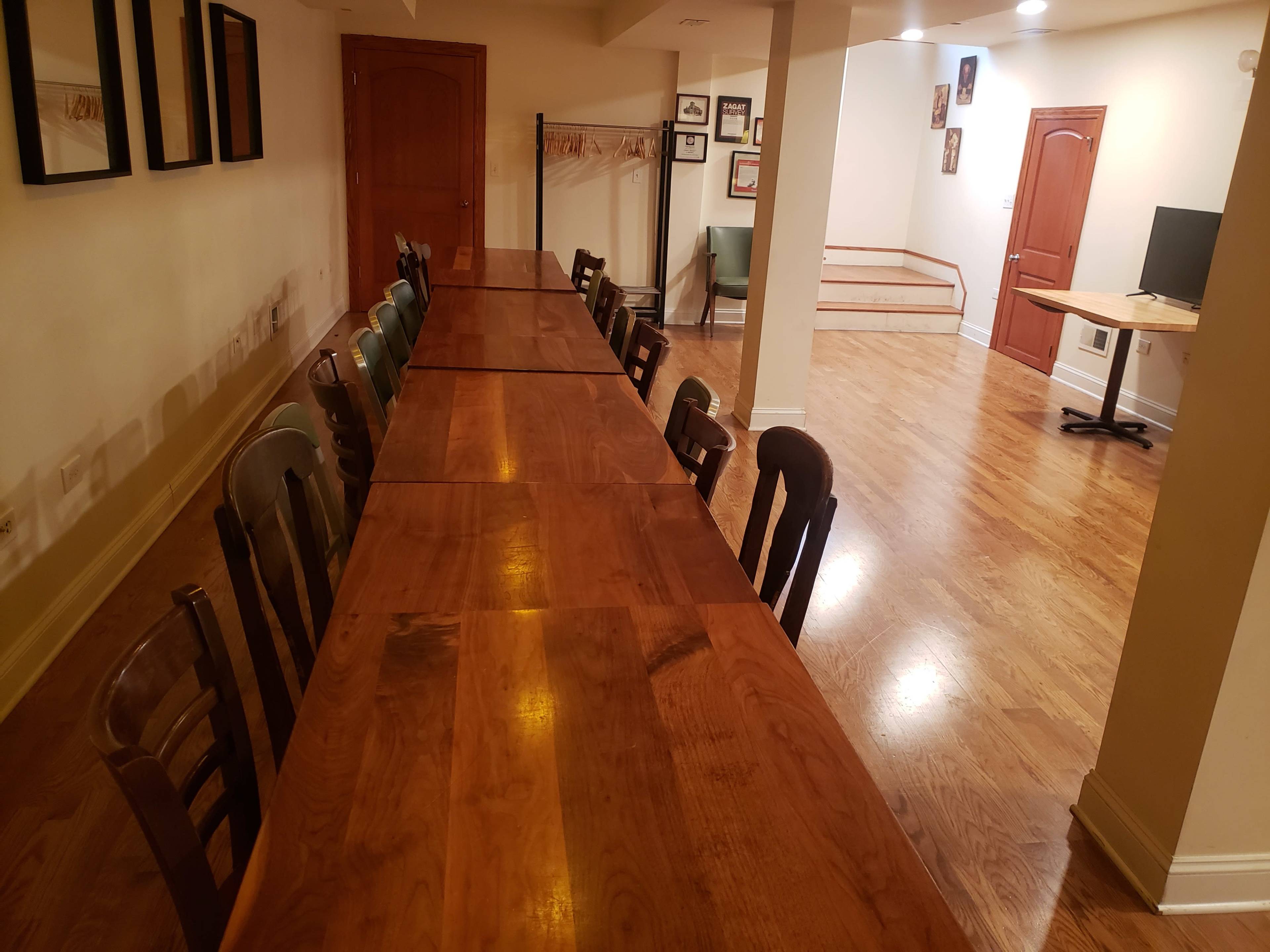 A large, empty room features a long wooden table with chairs on one side and a small seating area with a computer desk on the opposite side.