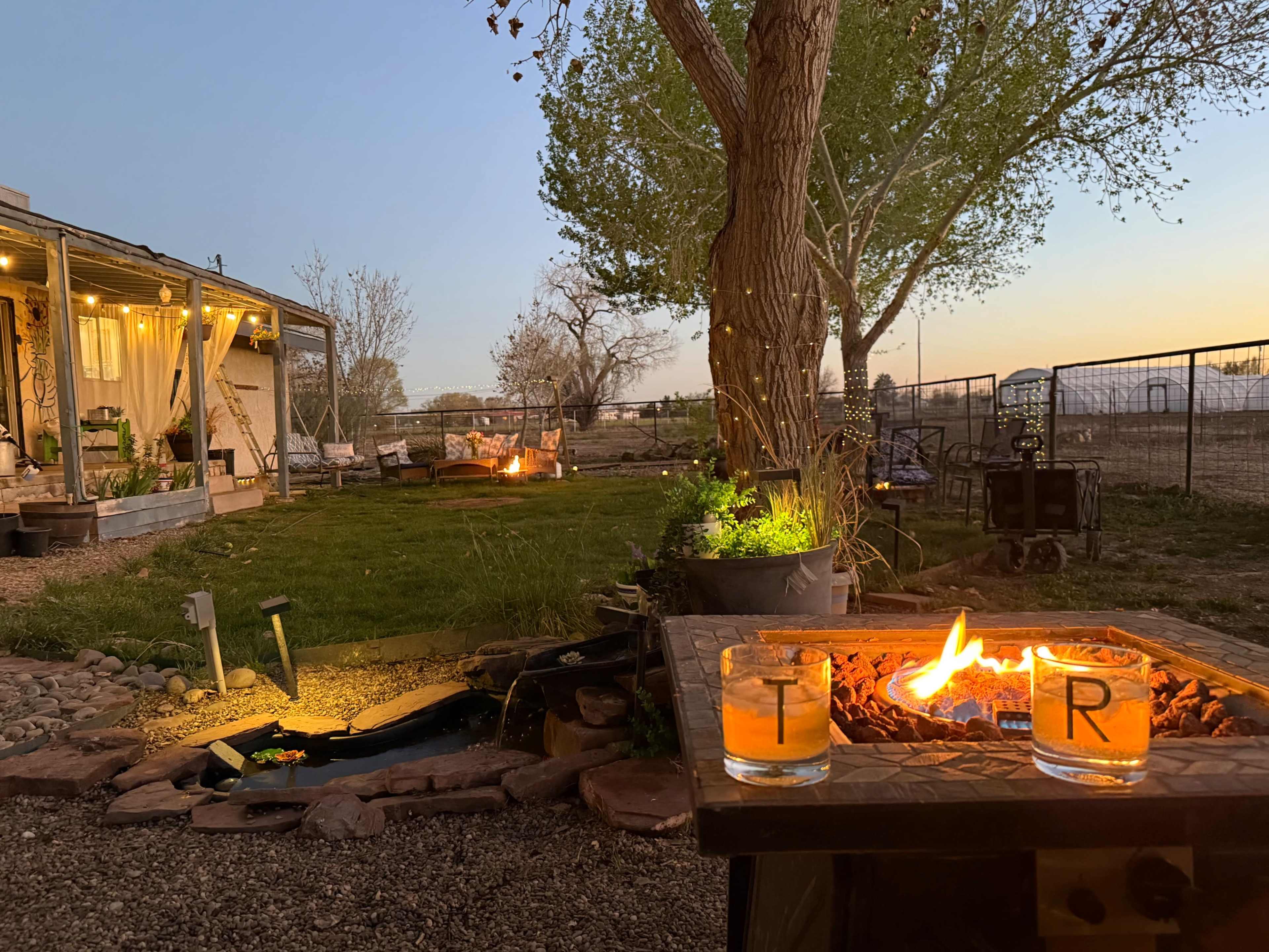 A backyard scene at dusk features a fire pit with two glasses resting on a tabletop, surrounded by lawn chairs and trees.