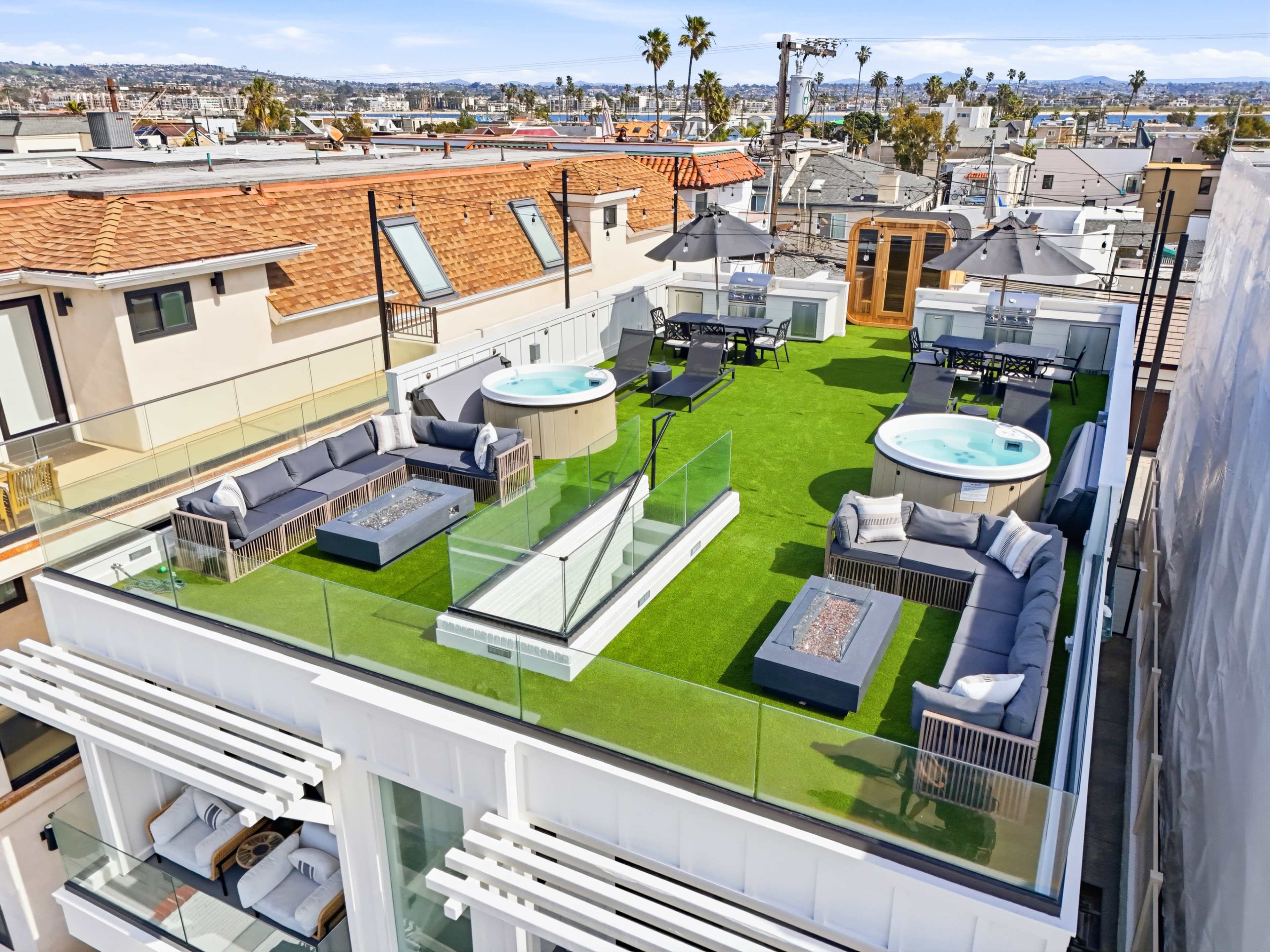 The image shows a rooftop terrace with two hot tubs, lounging furniture, and a grass-like surface, overlooking residential buildings and palm trees.