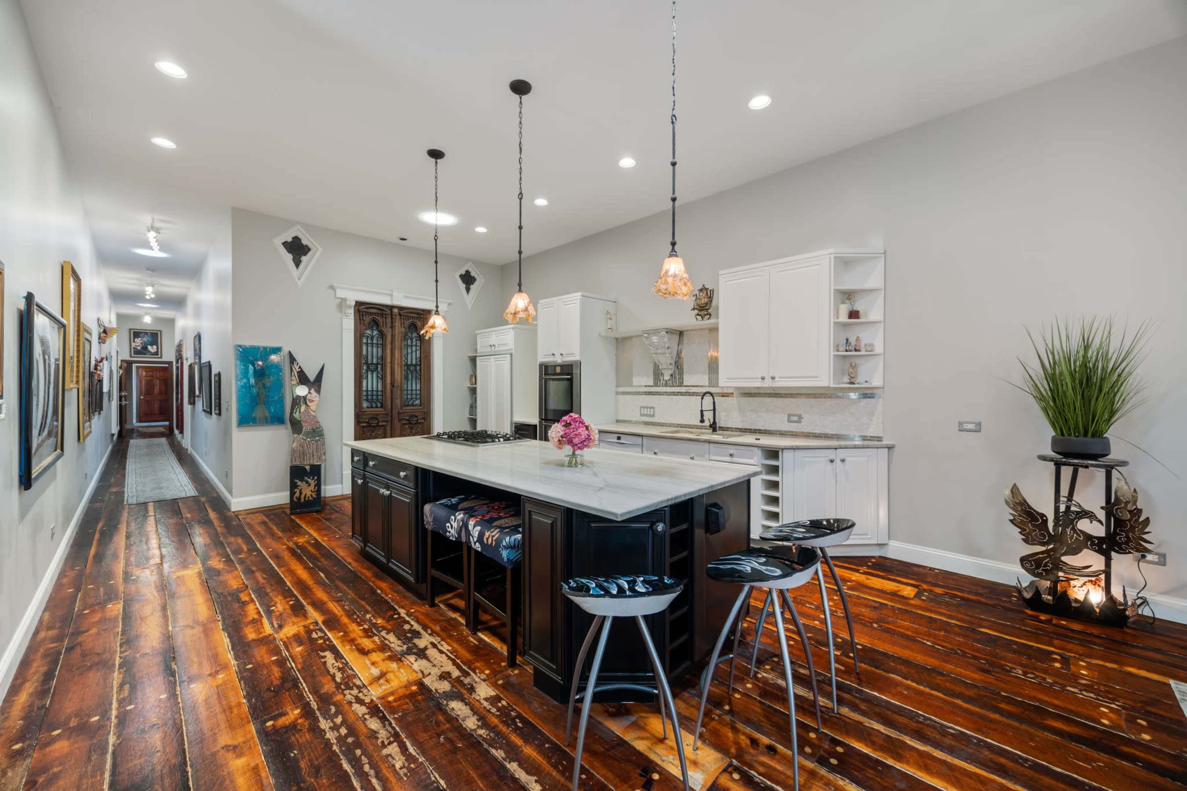 The image shows a modern kitchen with a large island featuring a marble countertop, black cabinetry, and pendant lighting, with rustic wooden flooring and a corridor lined with artwork in the background.