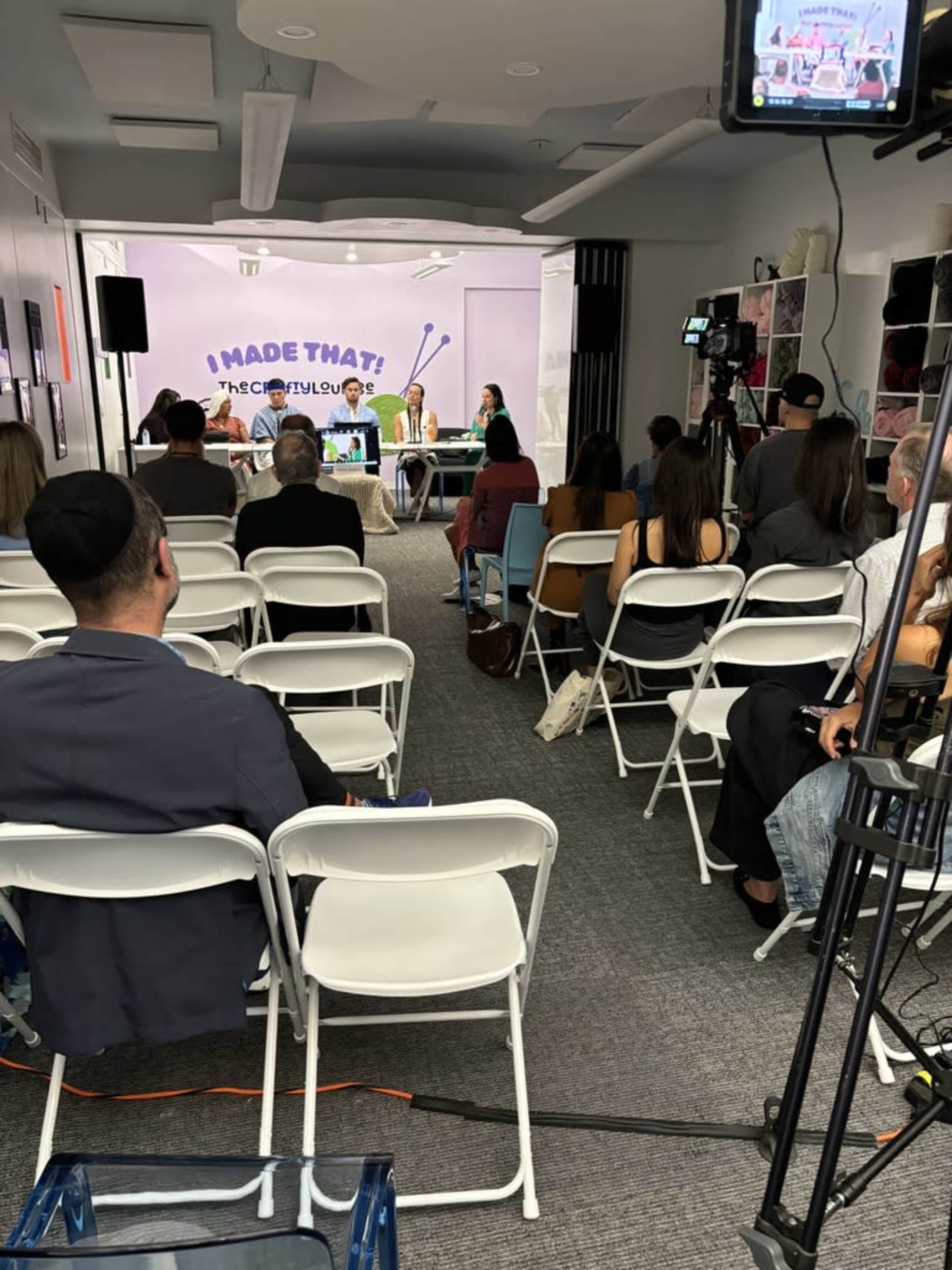 A panel discussion takes place in a room filled with seated audience members, while cameras and lighting equipment capture the event.