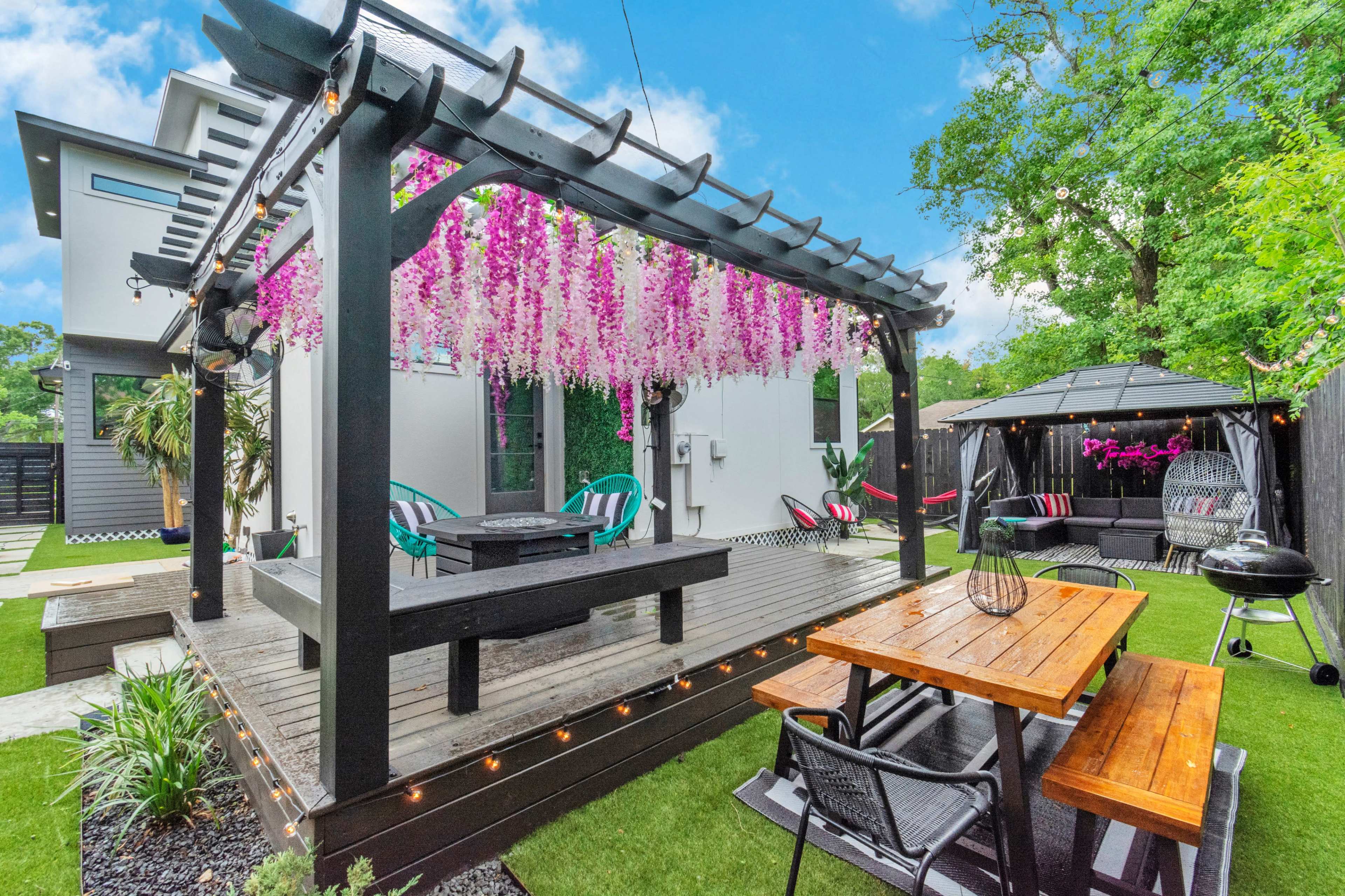 A landscaped backyard featuring a wooden deck with a pergola adorned with hanging purple flowers, surrounded by seating areas and a gazebo.