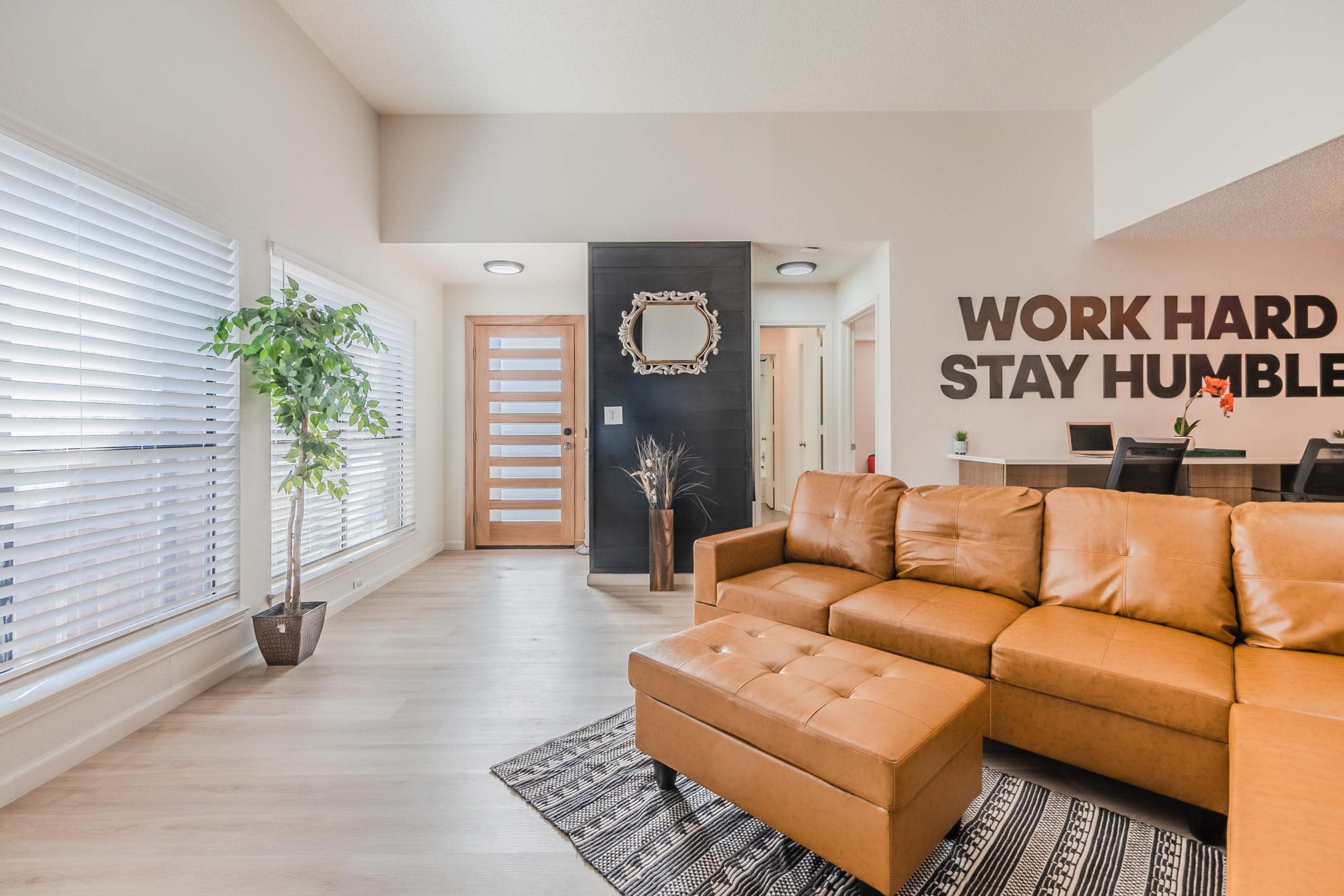 A modern living area features a brown leather sectional couch, a potted plant, large windows with blinds, and motivational wall decor that reads "WORK HARD STAY HUMBLE."