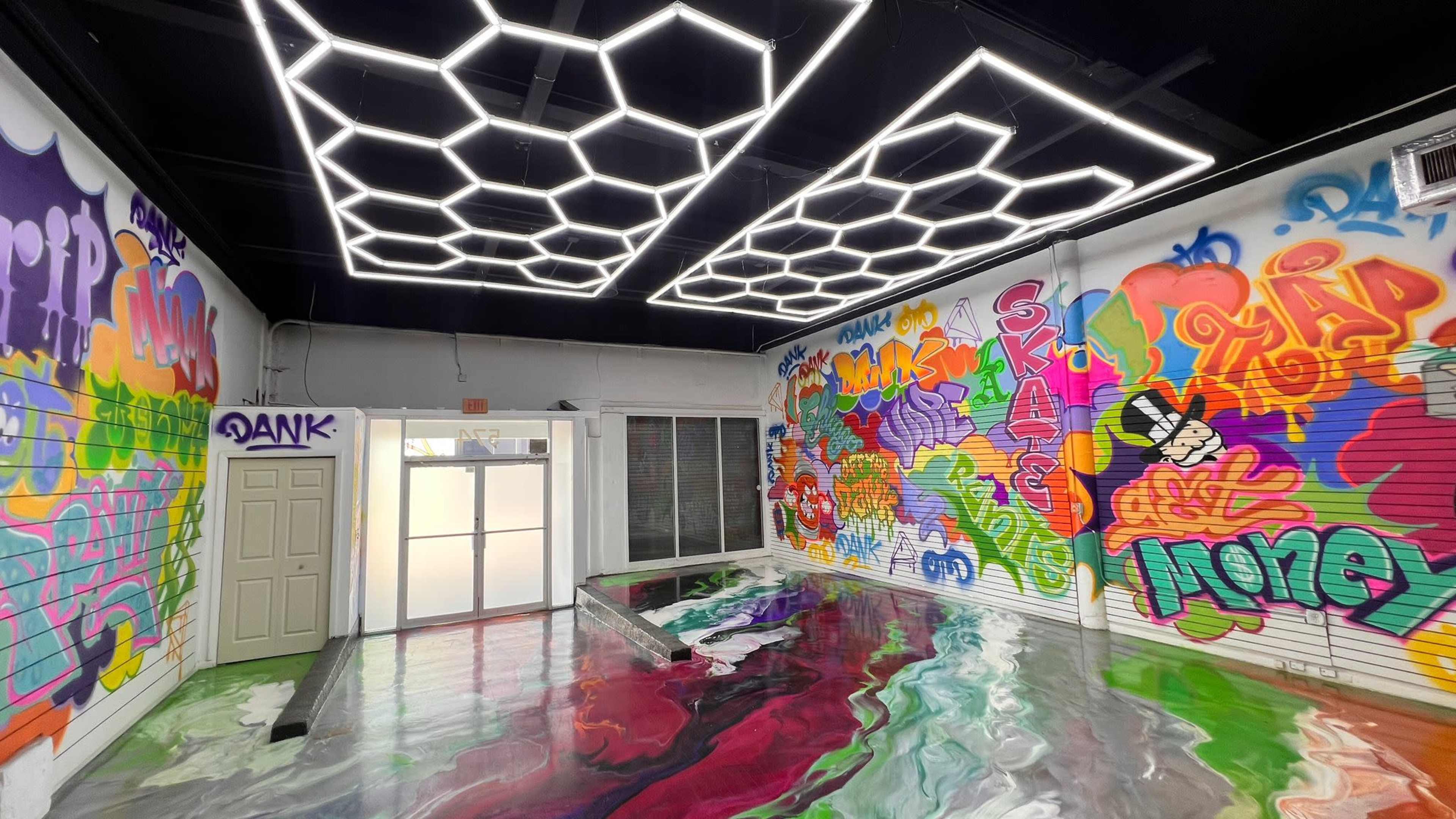 The image shows an indoor space featuring vibrant graffiti art on the walls and a glossy, colorful floor with a unique hexagonal light fixture overhead.