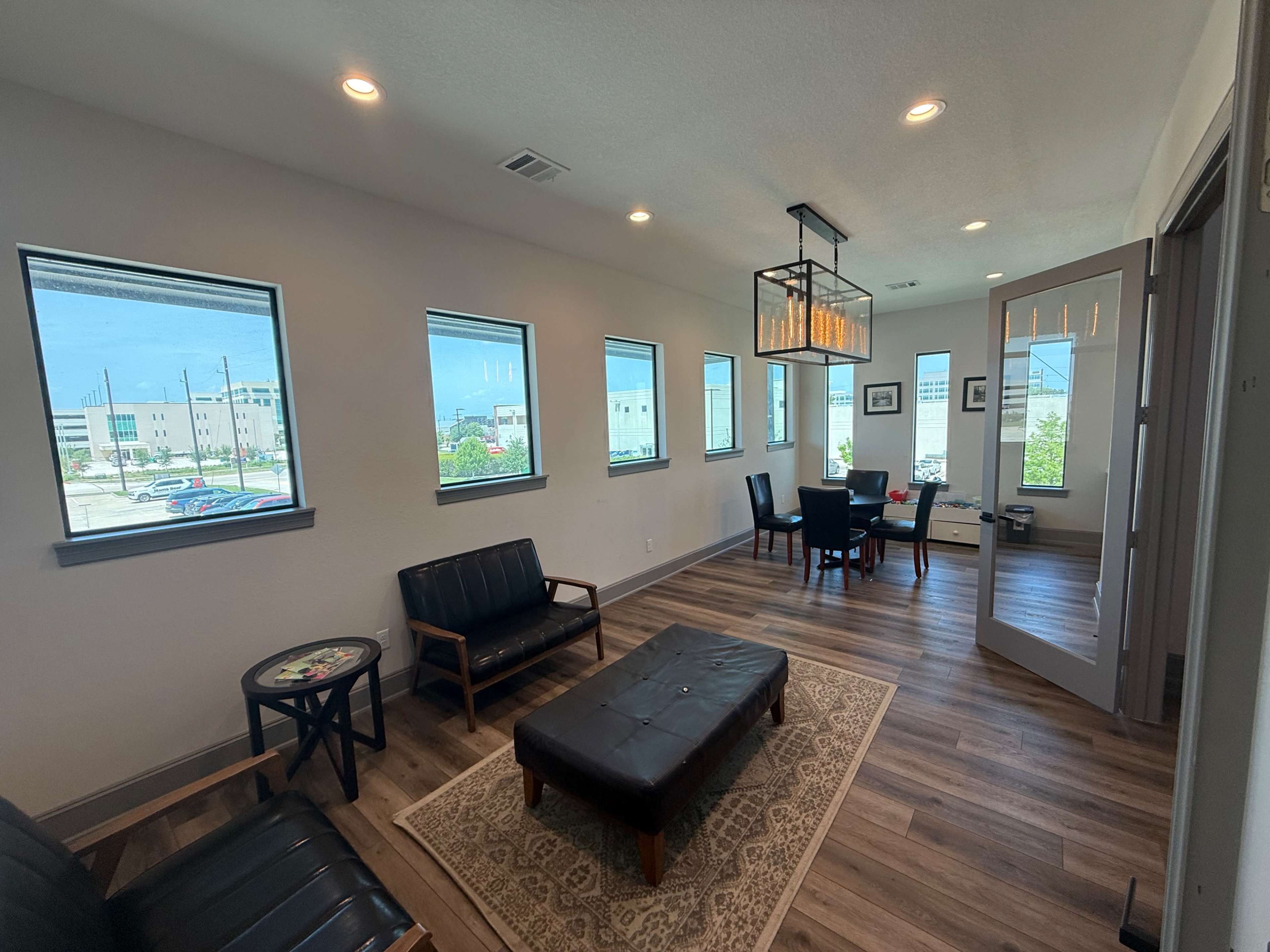 The image shows a modern, well-lit waiting area with two black sofas, a coffee table, and a dining area featuring a dark table and chairs.
