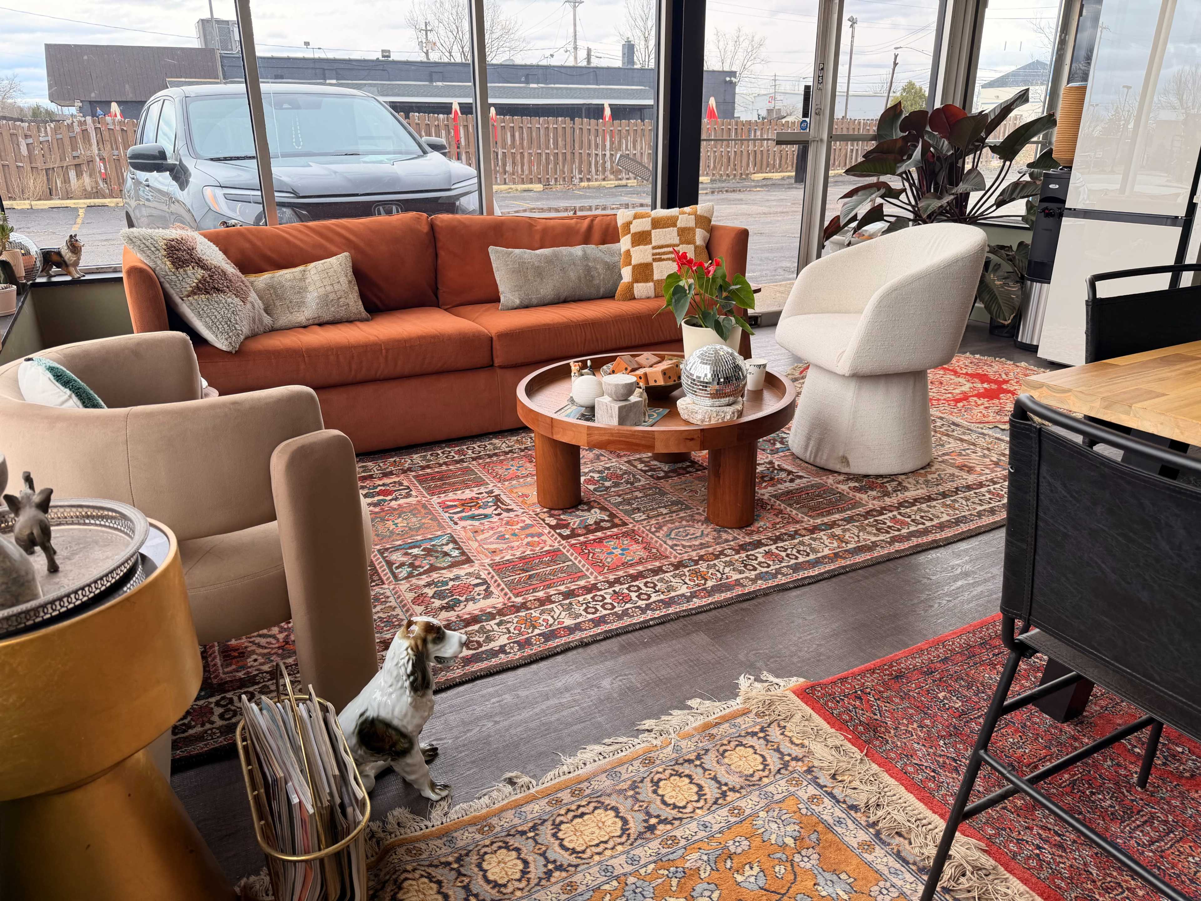 The image shows a modern living space with a large orange sofa, two armchairs, a round coffee table, and a decorative rug, all situated by large windows with a view of the outdoors.