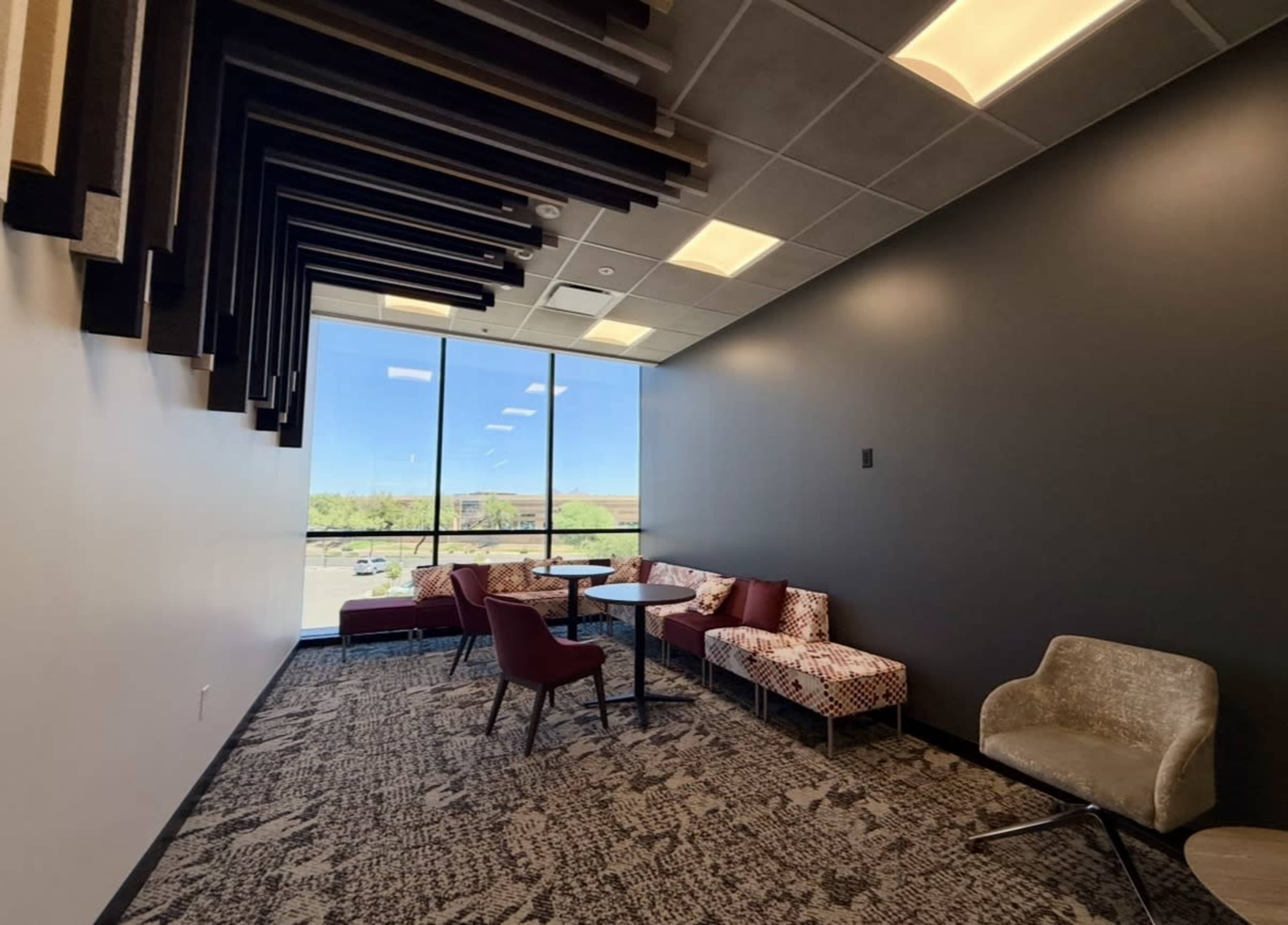 The image shows a modern lounge area with a mix of patterned and solid-colored seating arranged around a small table, situated near a large window.
