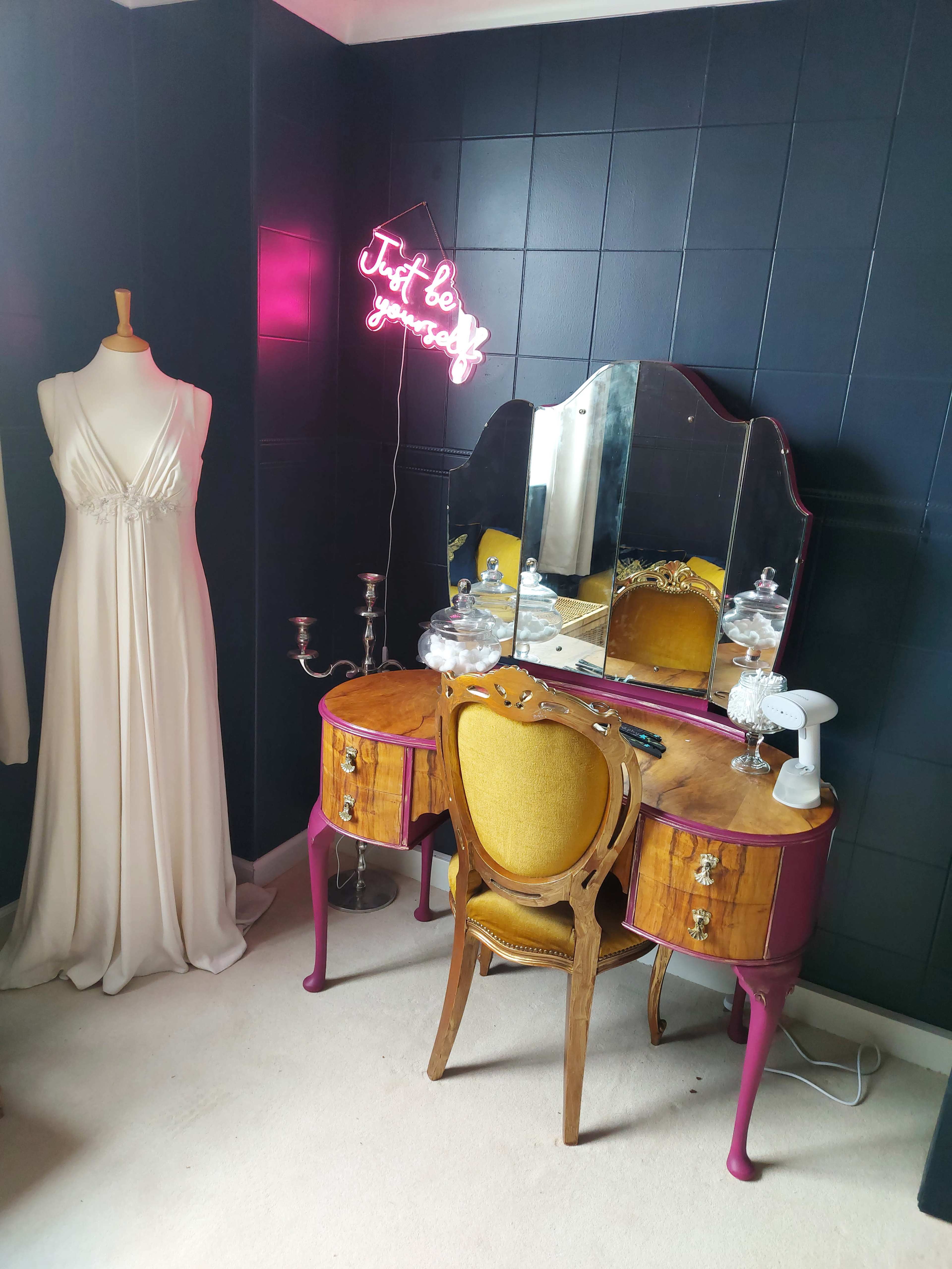 A vintage vanity table with a mirror and ornate chair is positioned beside a floor-length white dress and a neon sign that reads "Just be gorgeous."