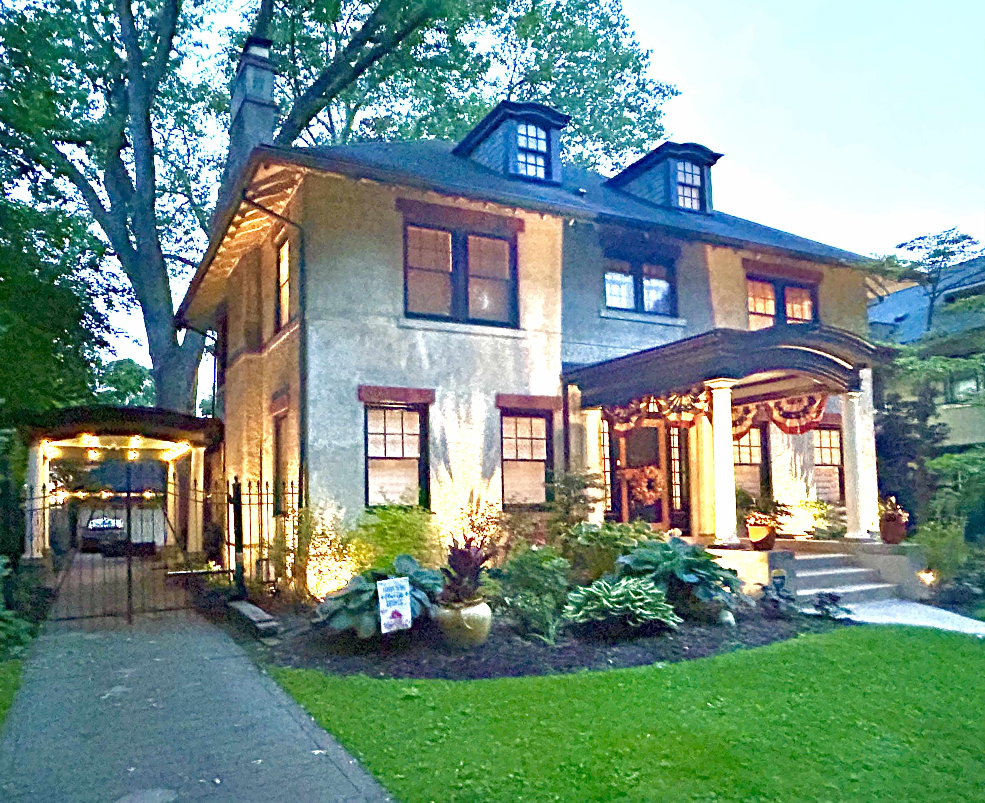 The image shows a two-story house with a spacious front porch, surrounded by a well-maintained lawn and garden, and a driveway leading to a garage.