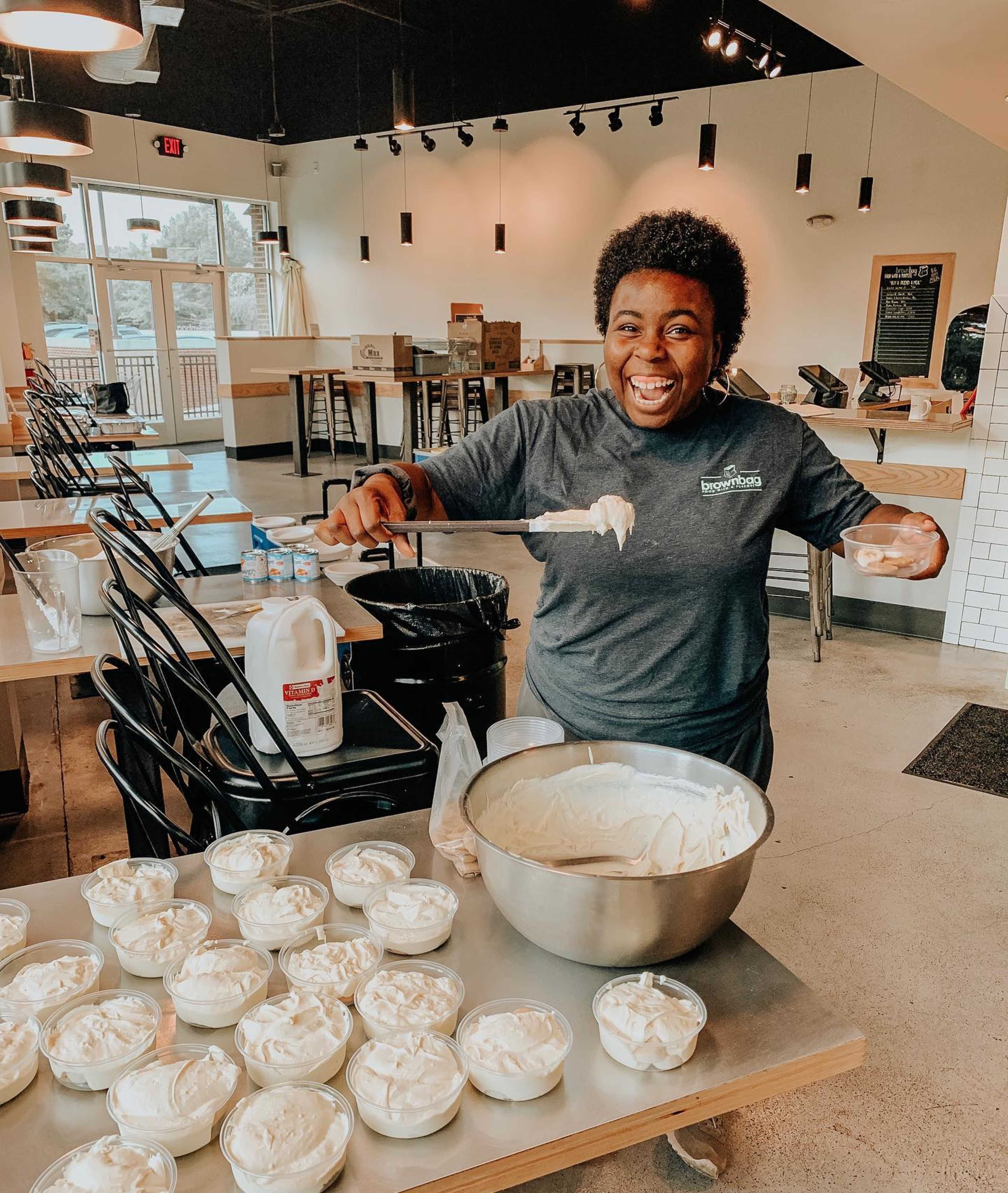 A person stands in a cafe, scooping ice cream from a large bowl into smaller containers on a table.