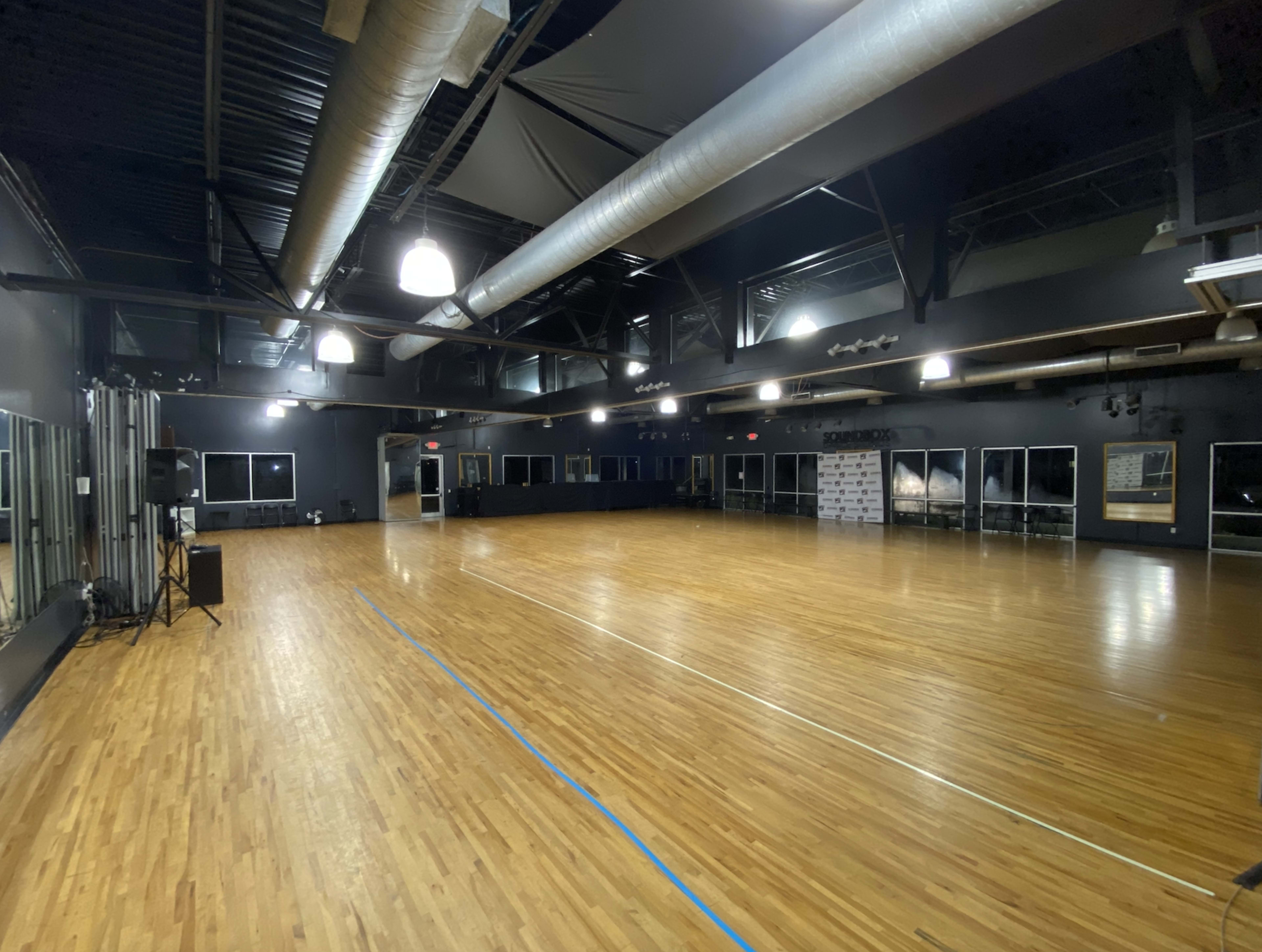 A spacious dance studio features a polished wooden floor, large mirrors, and high ceilings with industrial lighting.