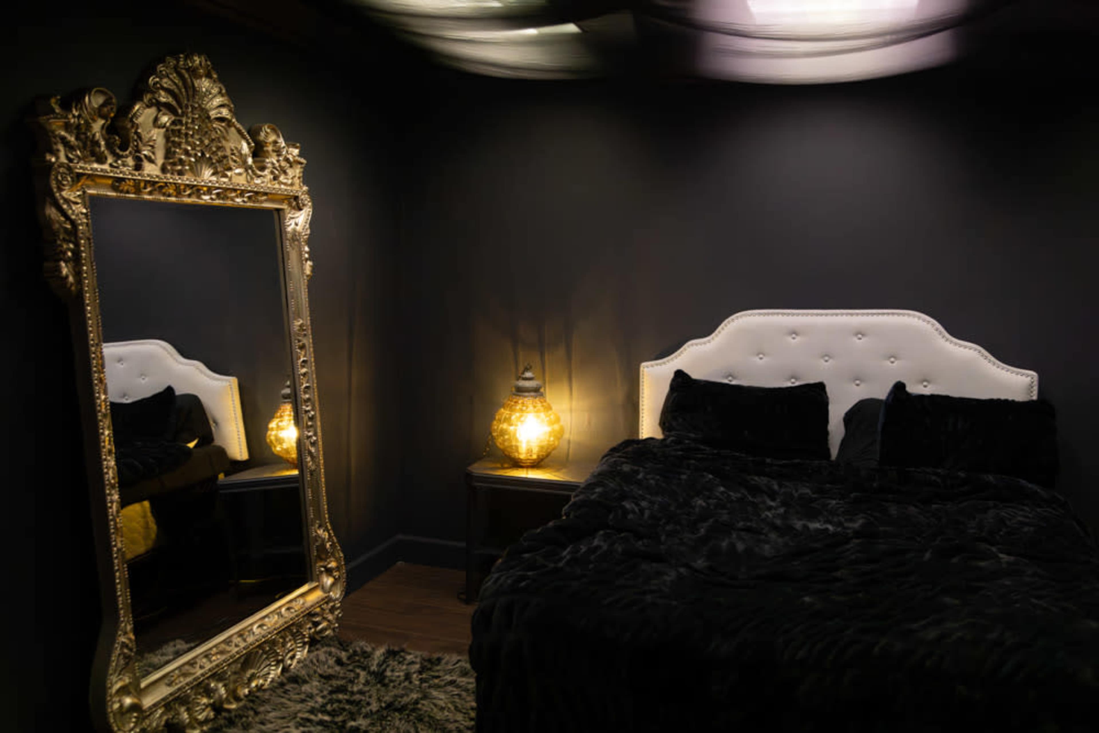 A dimly lit bedroom featuring a large ornate mirror, a white tufted bed with black bedding, and a decorative lamp on a nightstand.