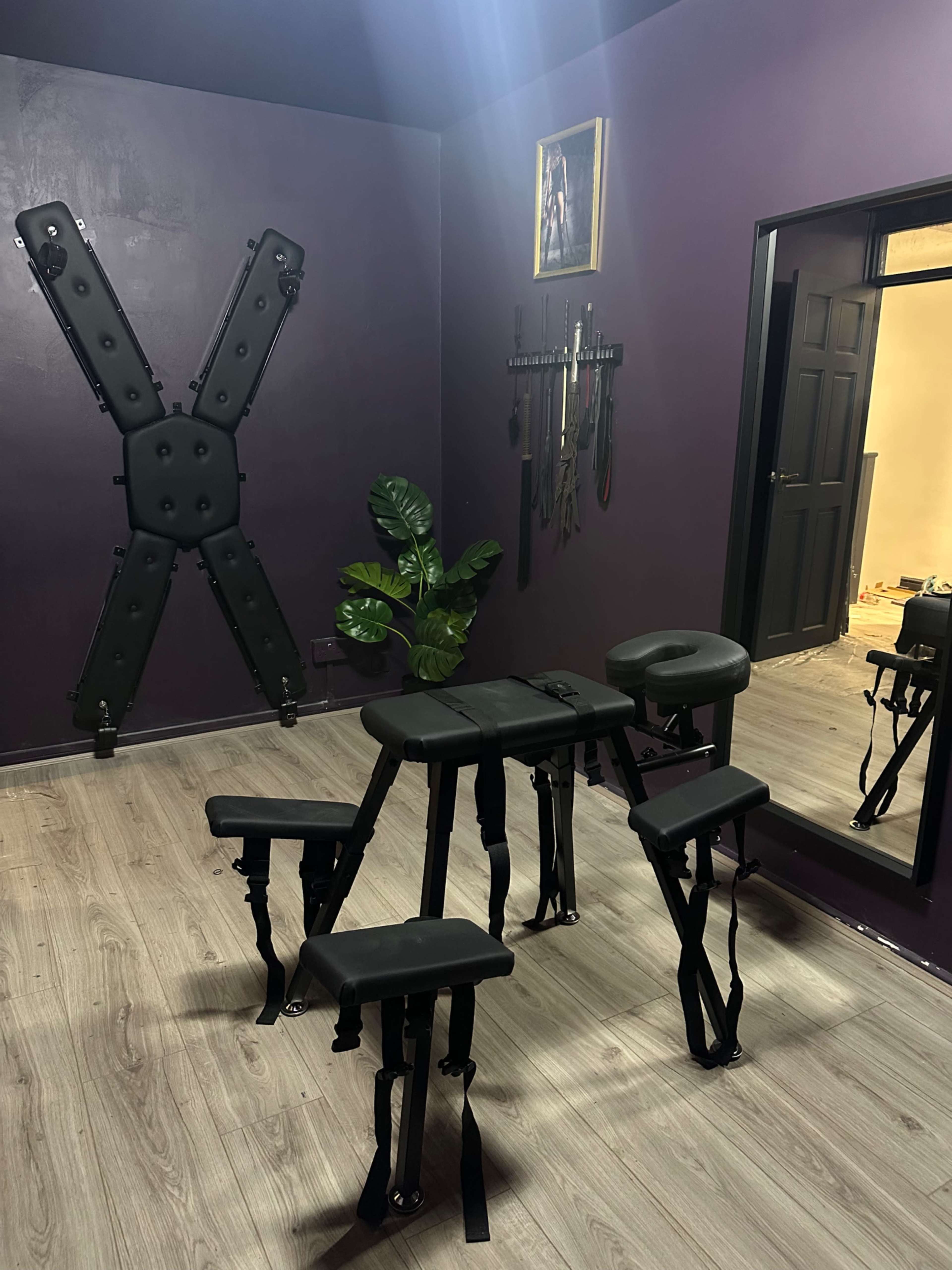 The image shows a dimly lit room with a dark purple wall, featuring various BDSM equipment, including a cross on the wall and a restraint table in the center.